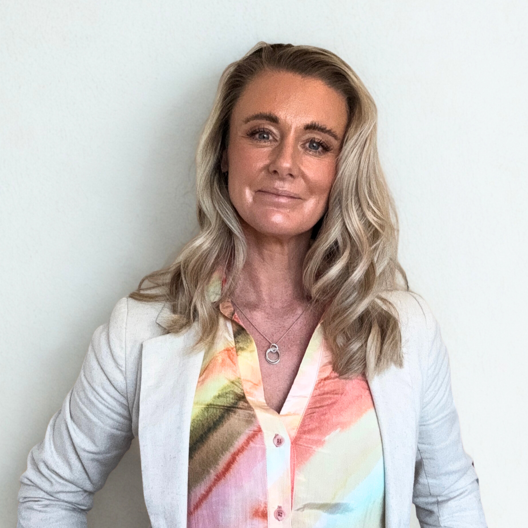 A woman with long, wavy blonde hair and light skin, wearing a white blazer over a colorful floral blouse, stands against a plain light-colored background.