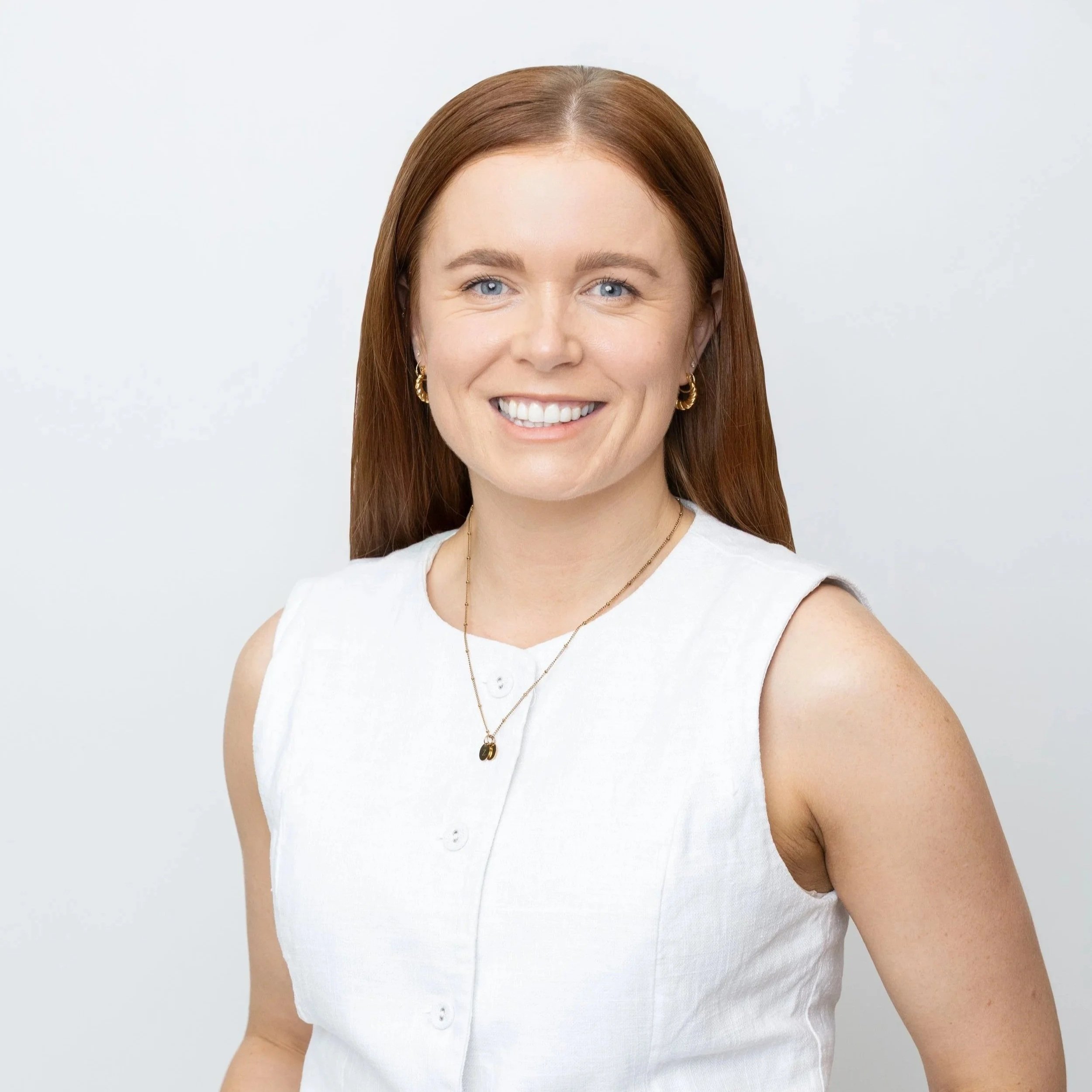 A woman with red hair, blue eyes, and earrings smiling, wearing a white sleeveless top and jewelry, standing against a plain light background.