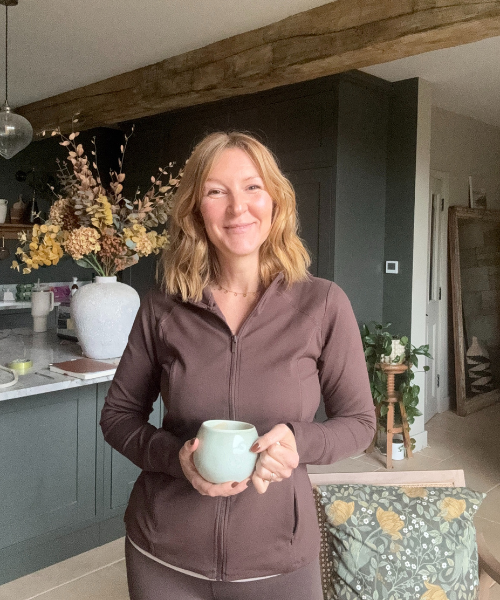 Lindsey Whitaker in a cozy, modern kitchen holding a green mug, wearing a relaxed plum-toned outfit. This calm, grounded moment reflects the spirit of her Midlife Reset program