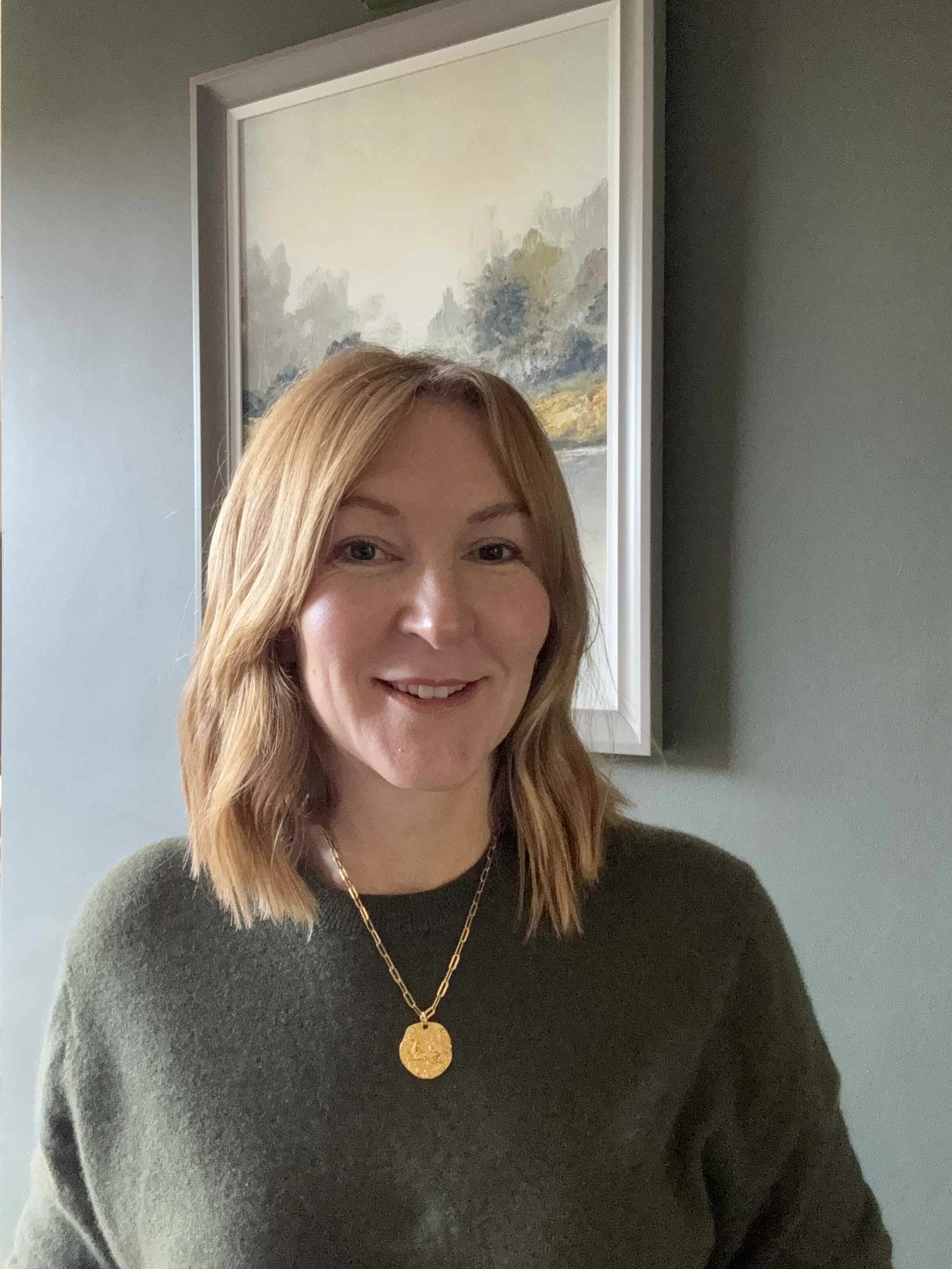 Lindsey Whitaker, Nutritional Therapist, Nurse, and Personal Trainer, standing calmly indoors in front of a misty landscape painting. She wears a dark green sweater and a gold pendant.