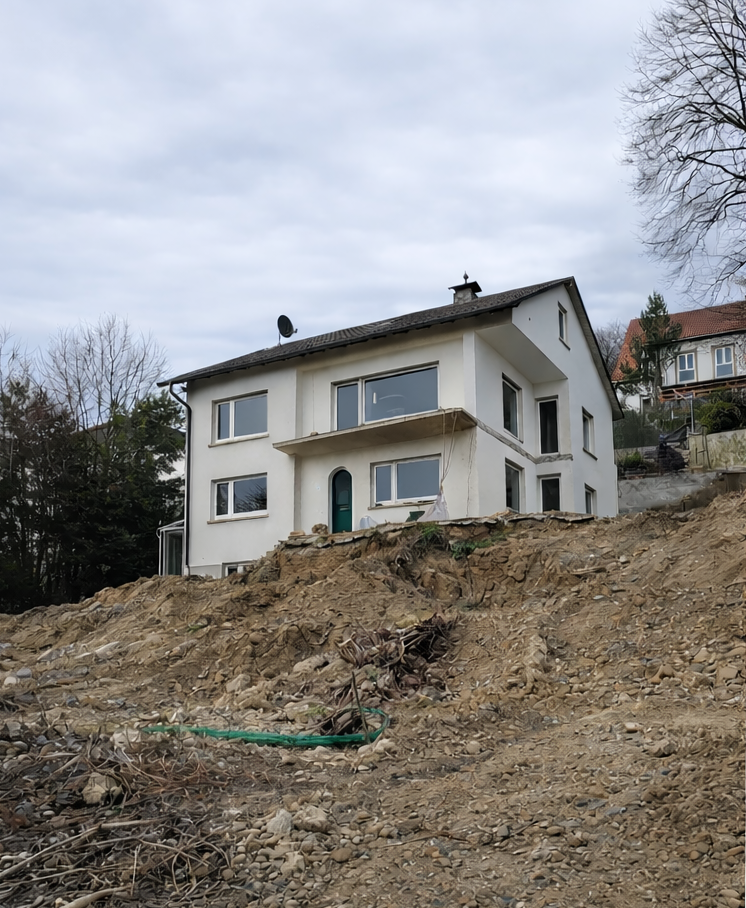 Ein mehrstöckiges weißes Haus auf einem Abhang, umgeben von Bäumen, im Hintergrund weiterer Häuser, der Boden ist aufgerissen und vereist.