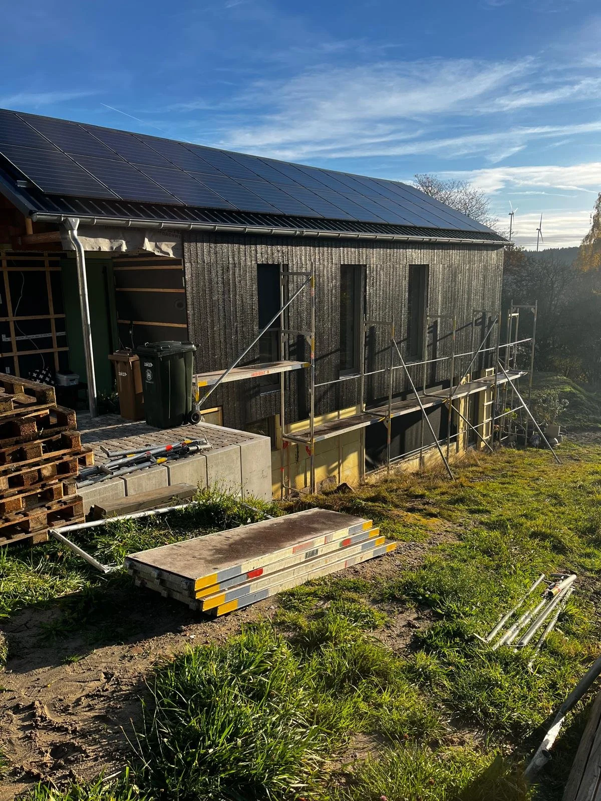Baustelle an einem modernen Haus mit Solardächern, im Vordergrund liegen Baugerüste, Holzkisten und Baumaterialien.