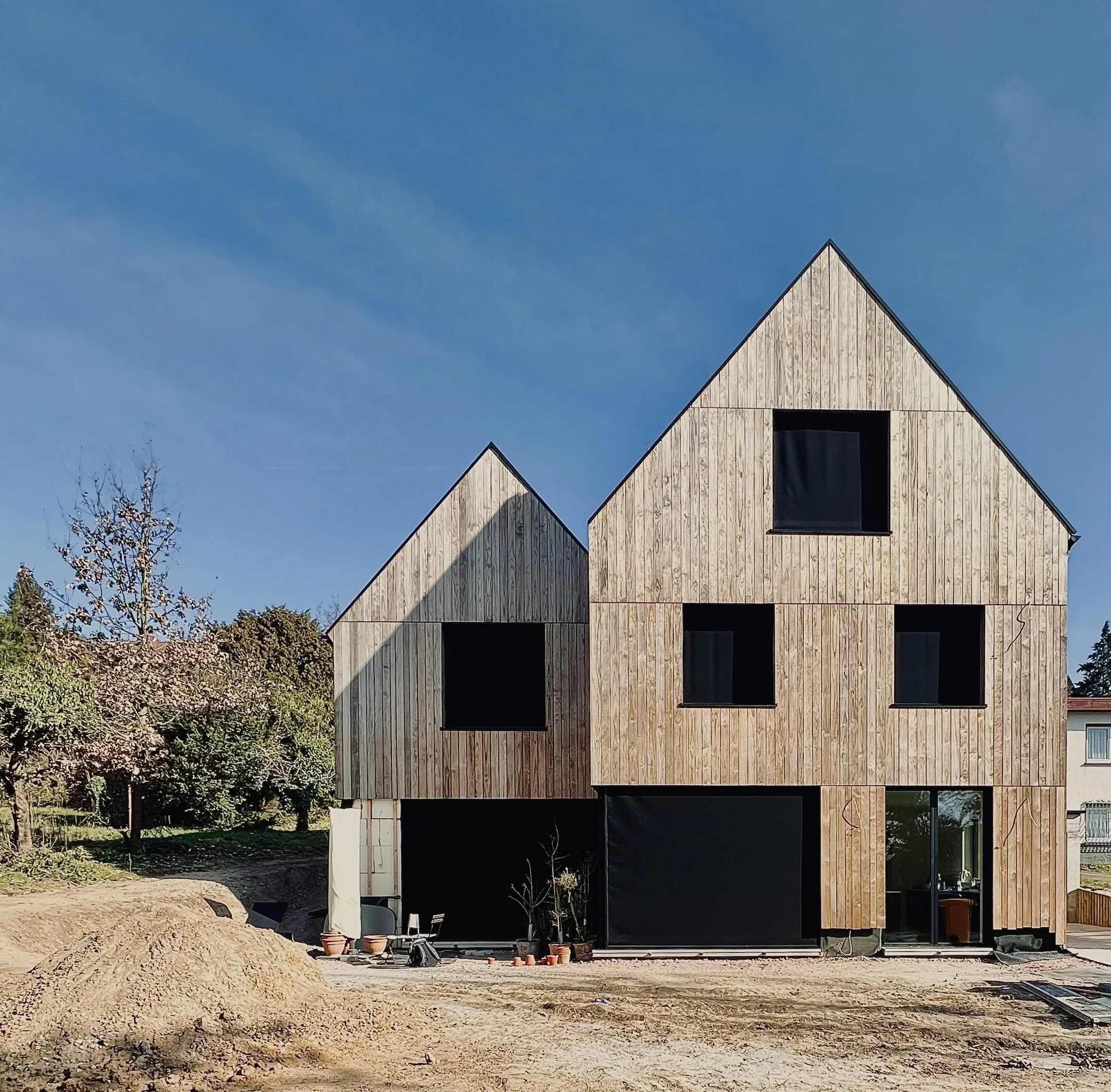 Neues Haus mit Holzfassade und schwarzen Fenstern unter blauem Himmel, Baustelle im Vordergrund.