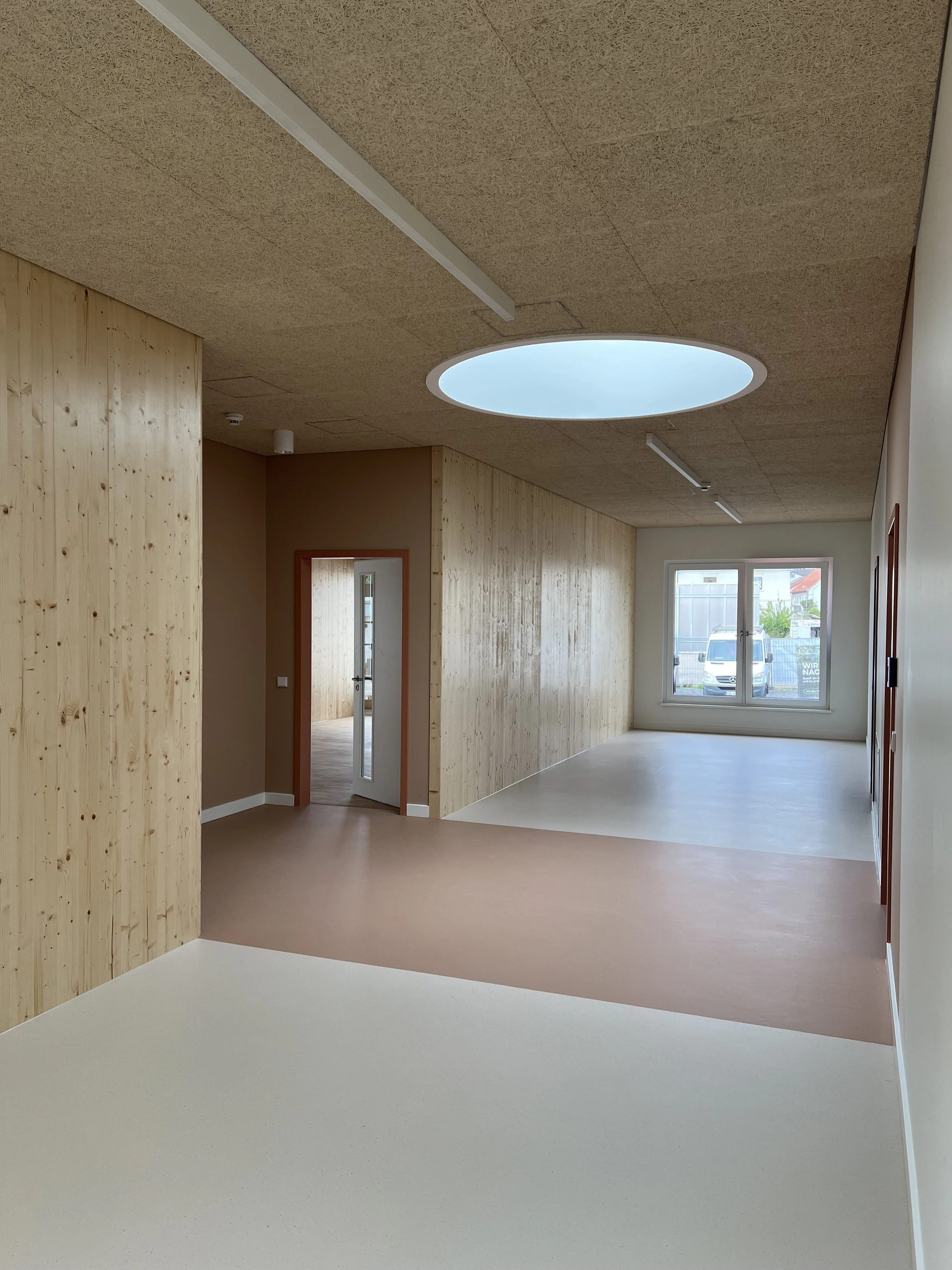 Innenraum eines modernen, lichtdurchfluteten Raums mit Holzpaneelen an den Wänden, großen Fenstern, einem runden Oberlicht in der Decke und verschiedenen Bodenbelägen.