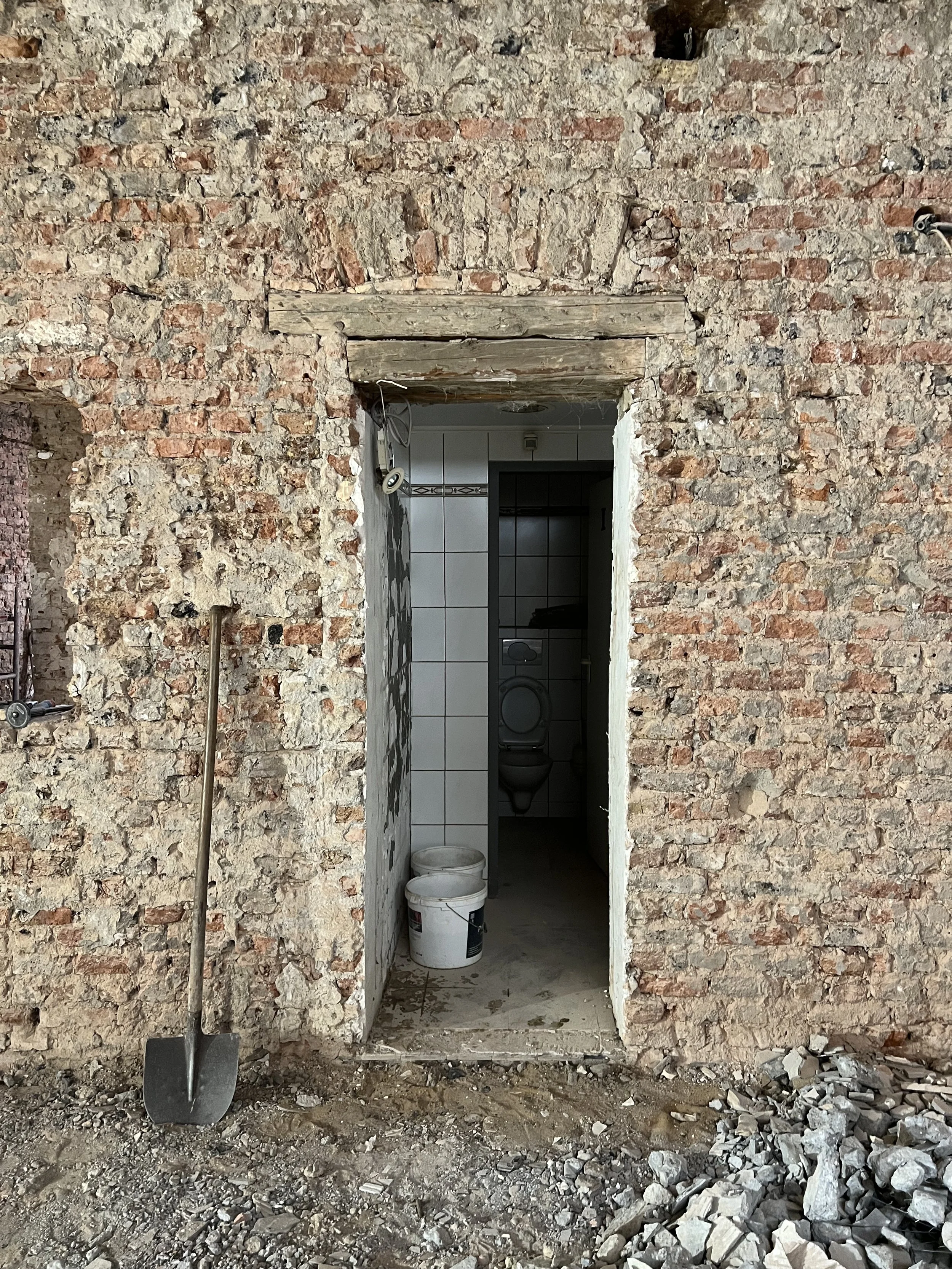 Baustelleneingang mit rotem Backsteinmauer, im Inneren ein WC mit Türrand, Putzwaren und einem Werkzeug im Vordergrund, im Vergleich zu einem unbefestigten Boden mit Bauabfällen.