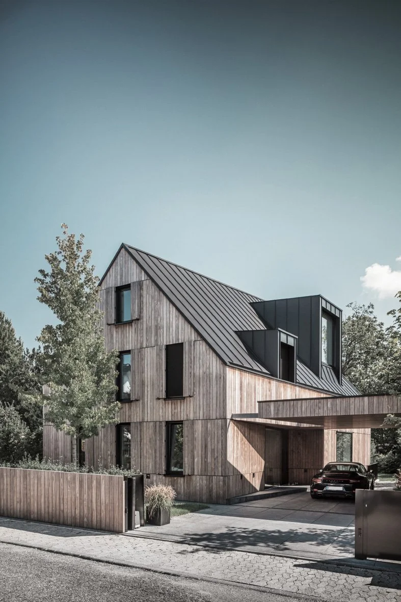 Ein modernes Haus mit einer Holzfassade, schräg geneigtem Dach und großen Fenstern, umgeben von Bäumen und einem Garten, mit einem Carport und einem schwarzen Auto davor.