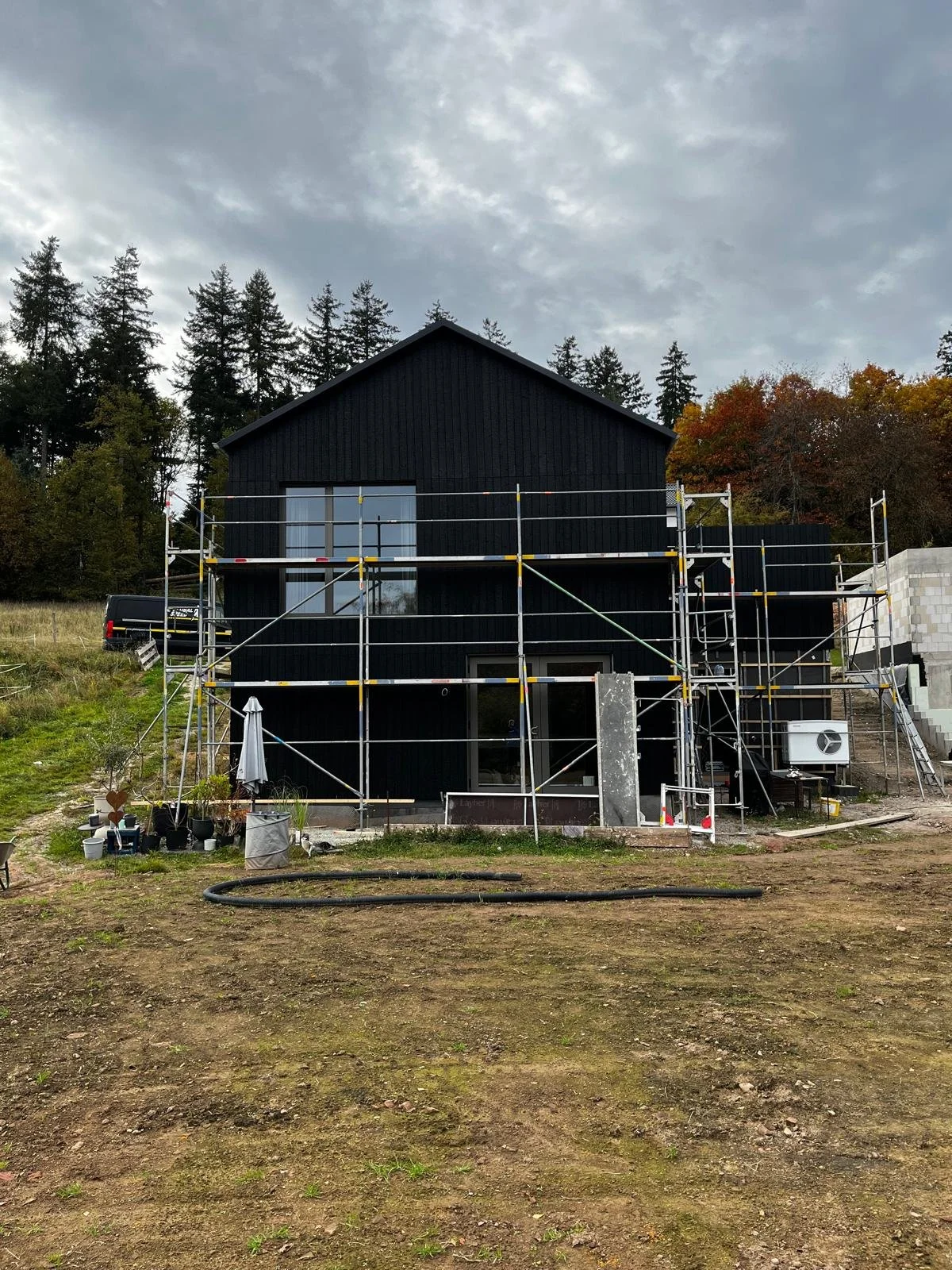 Ein schwarzes Haus im Bau mit Gerüsten umgeben und einem Himmel mit Wolken im Hintergrund, umgeben von Bäumen und Kiesboden im Vordergrund.