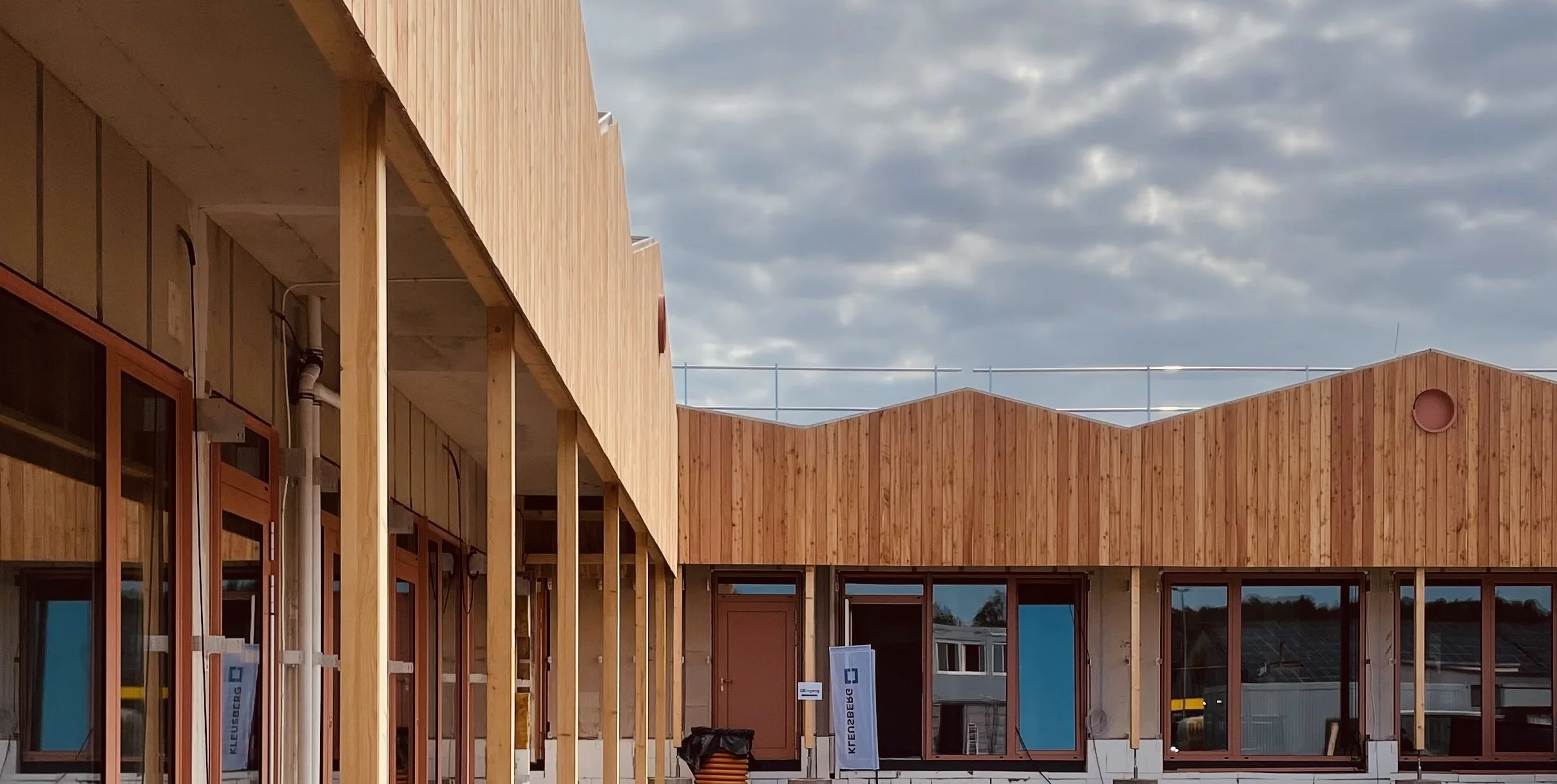 Neubau eines Gebäudes mit Holzfassade und großen Fenstern, Baugerüst im Hintergrund, bewölkter Himmel.