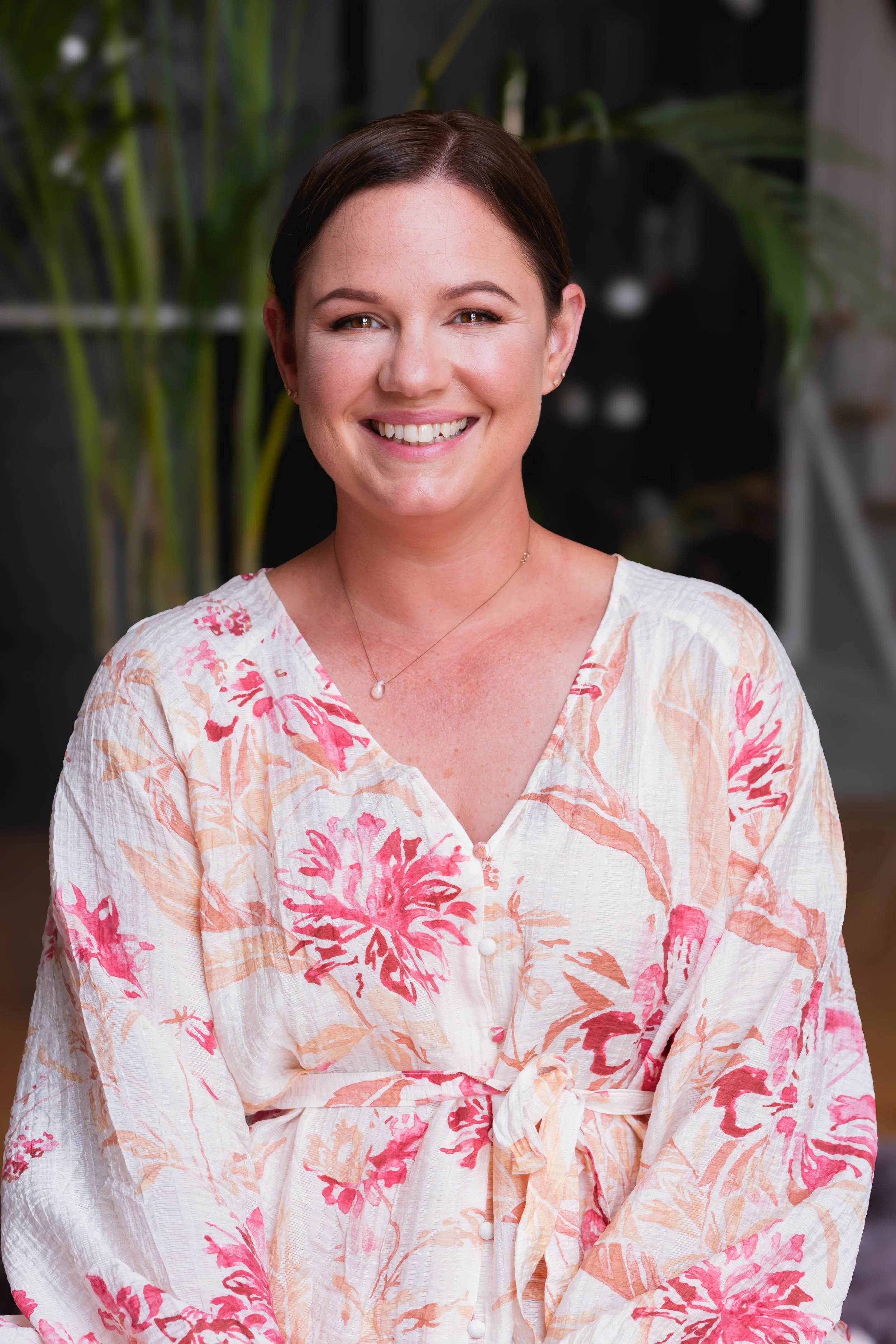 A smiling woman with short brown hair, wearing a floral blouse with pink and orange flowers, a pearl necklace, and small earrings, standing indoors with green plants in the background.