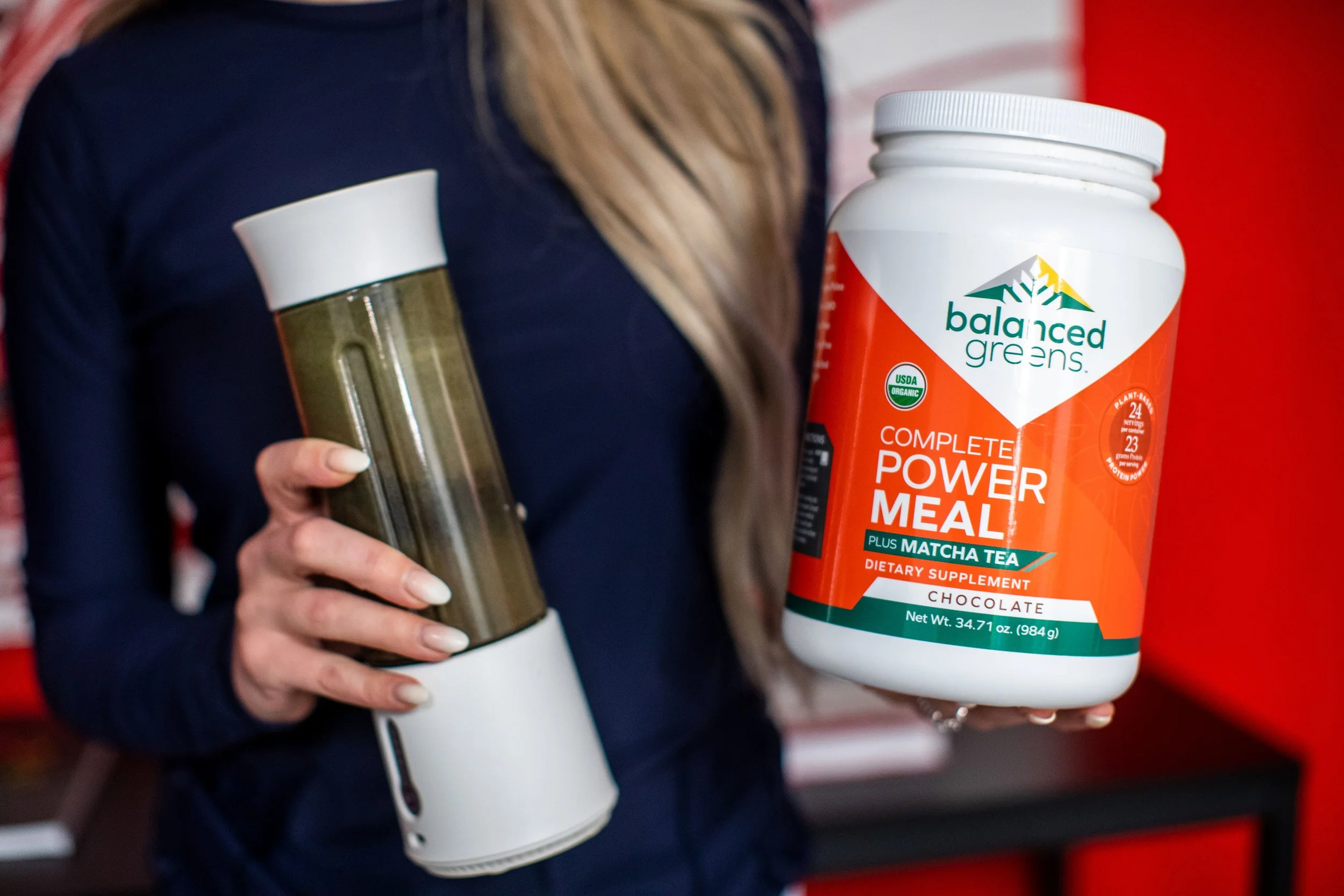 Person holding a green smoothie bottle in one hand and a container of Balanced Greens Complete Power Meal with Matcha Tea in the other, standing in front of a red background.