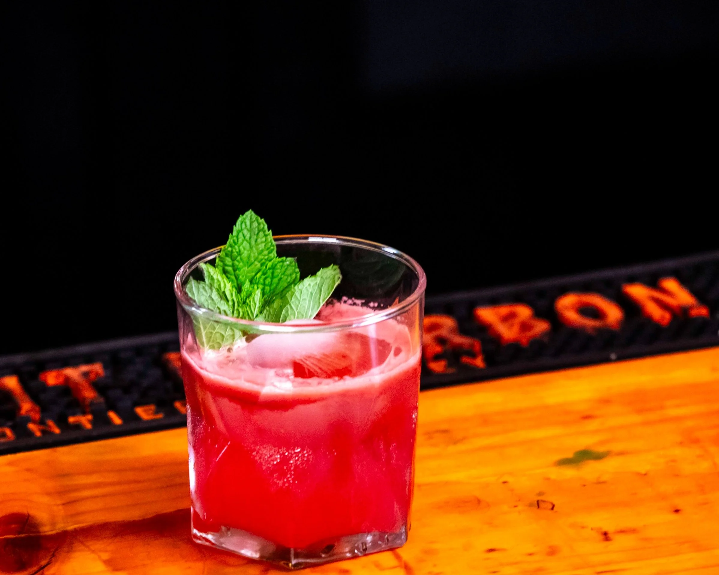 Red cocktail in a glass garnished with fresh mint leaves on a wooden bar counter.