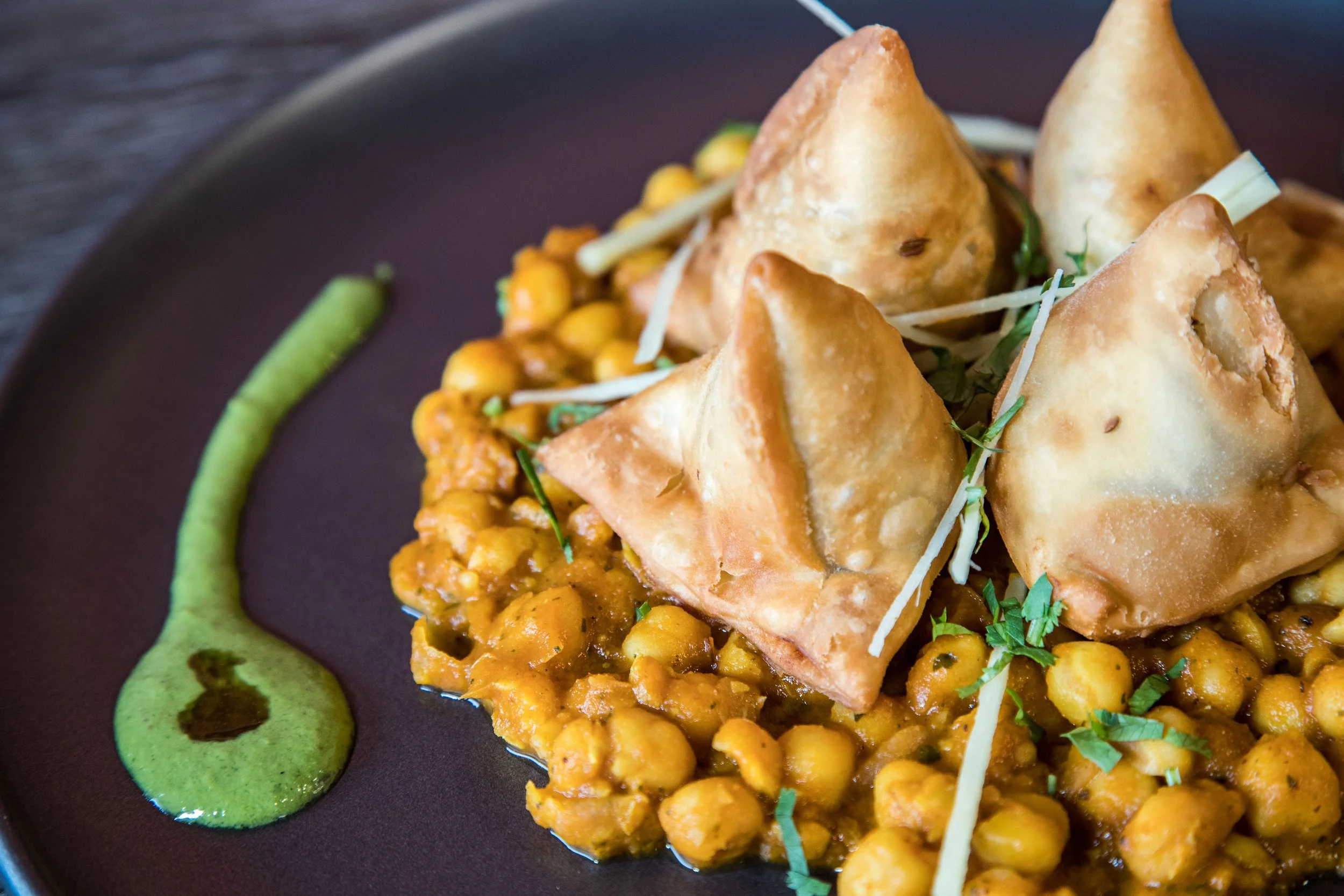 Indian dish with samosas, chickpea curry, garnished with herbs, and green sauce on a dark plate.