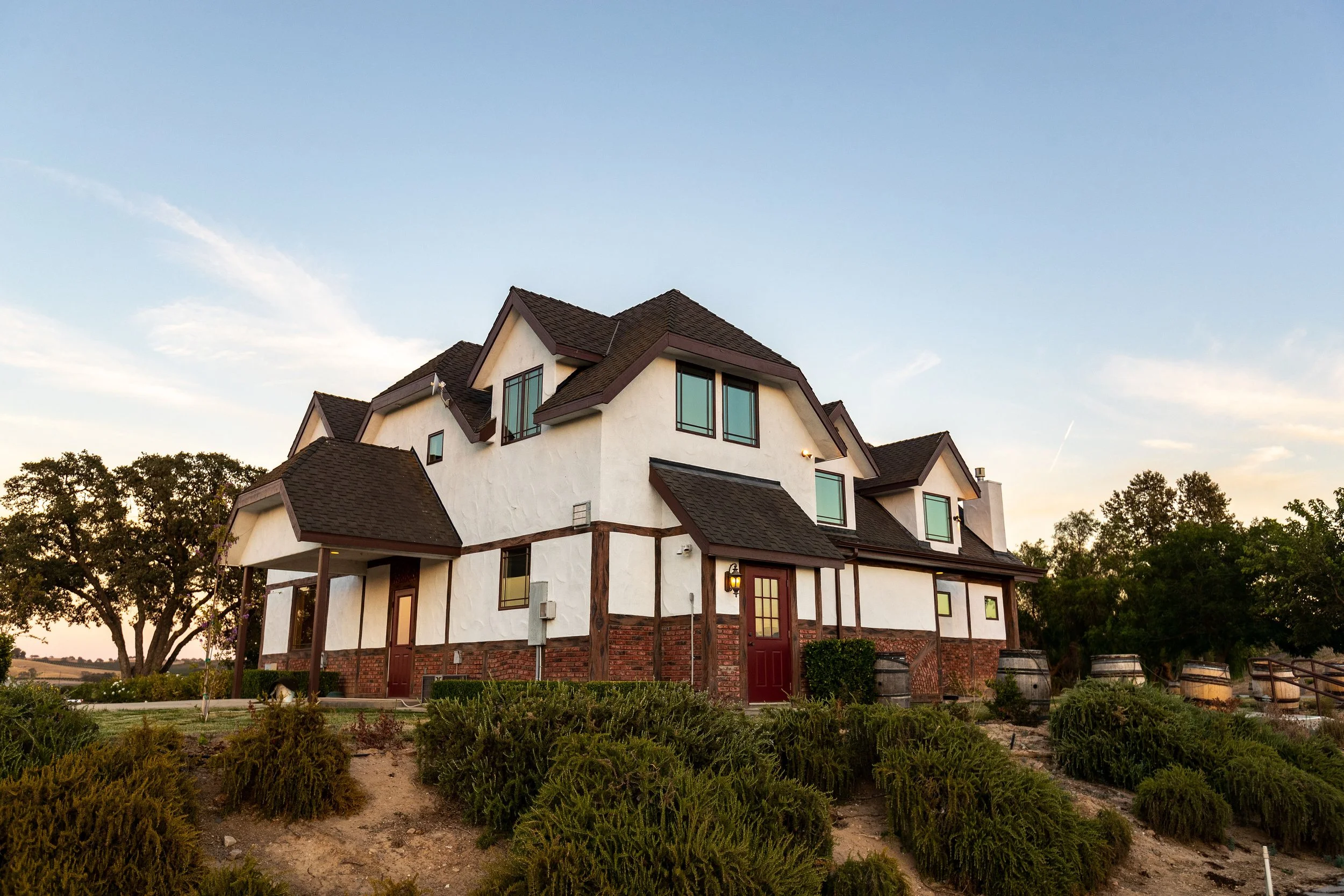 A large, multi-story house with white walls, brown roof, and brick accents, surrounded by bushes and trees, under a clear sky during sunset.