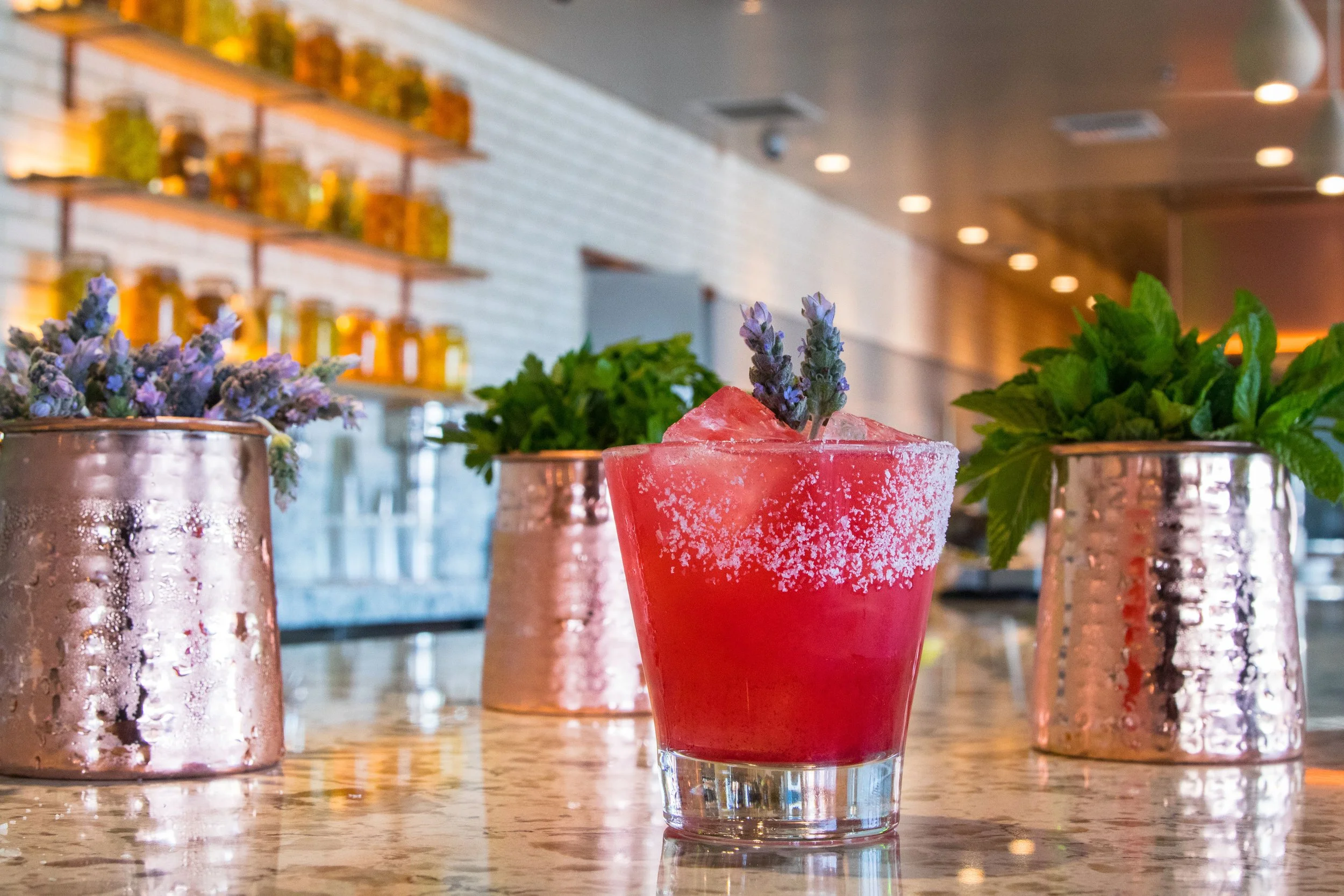 A red cocktail garnished with a sprig of lavender, placed on a marble countertop with two pots of lavender and mint plants in the background.