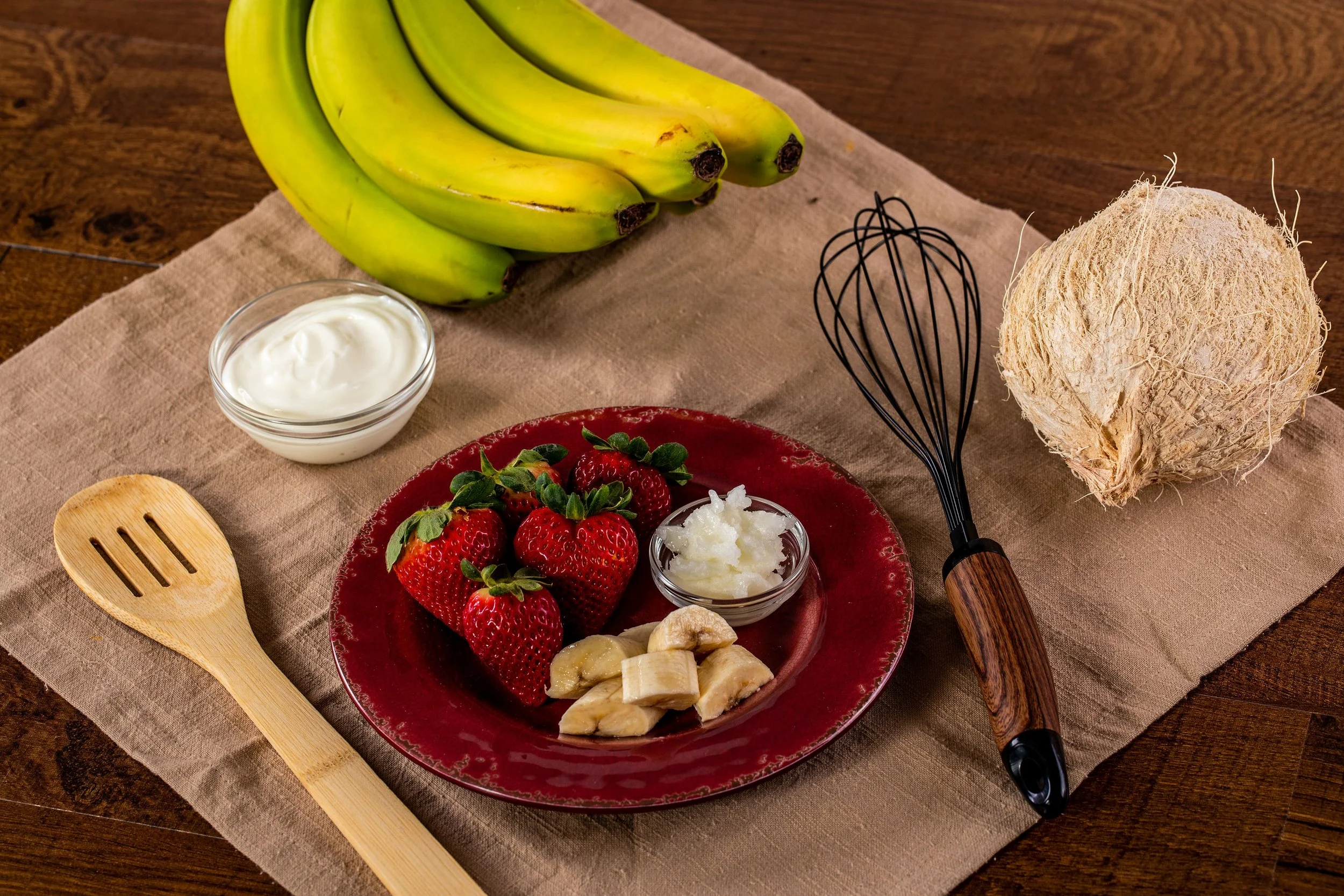 Bunch of bananas, bowl of yogurt, strawberries, banana slices, coconut, whisk, wooden spatula, and a bowl of coconut oil on a brown table with a beige cloth.