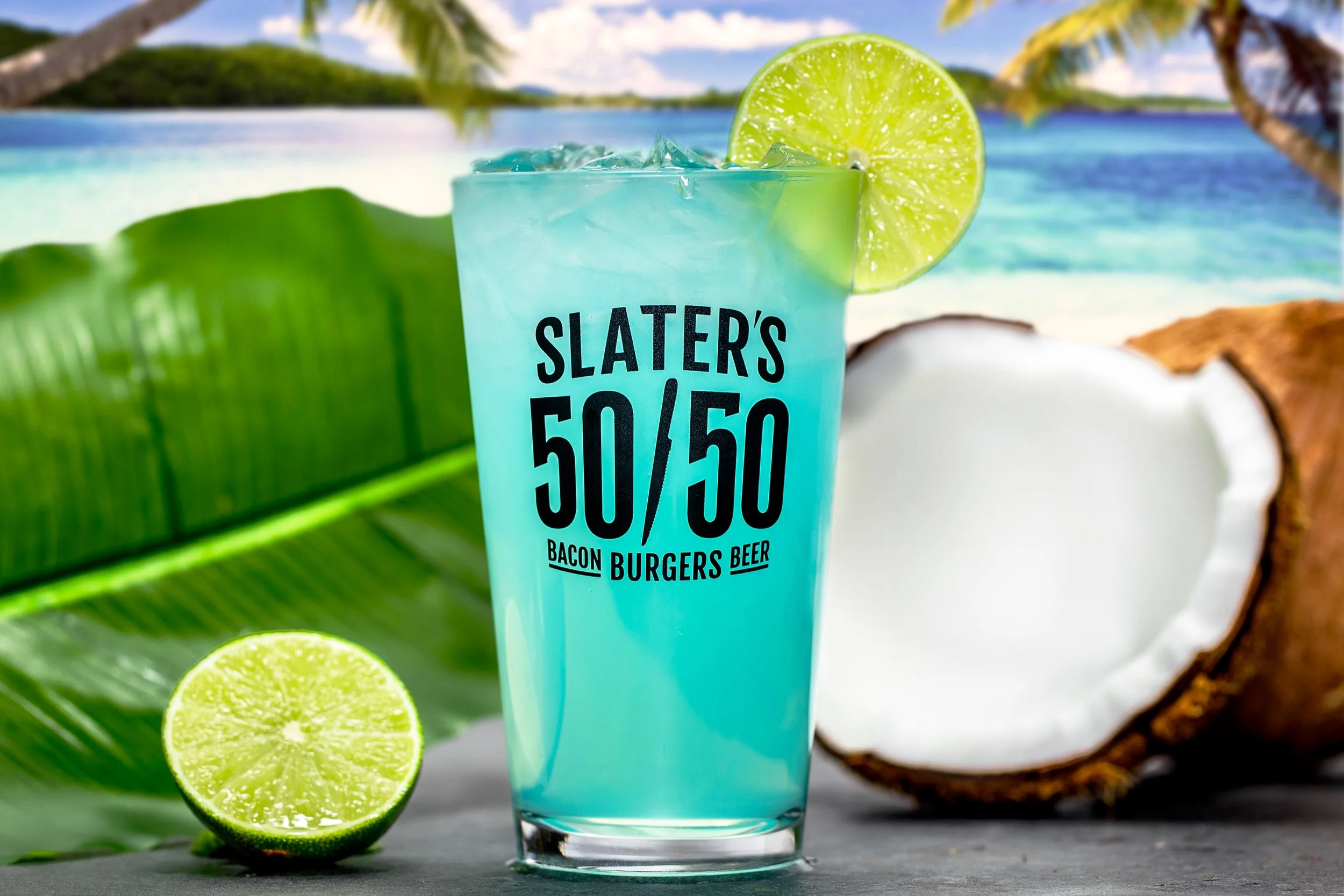 A blue tropical cocktail garnished with a lime wedge, with a coconut and a lime next to it, set against a beach background with palm trees.
