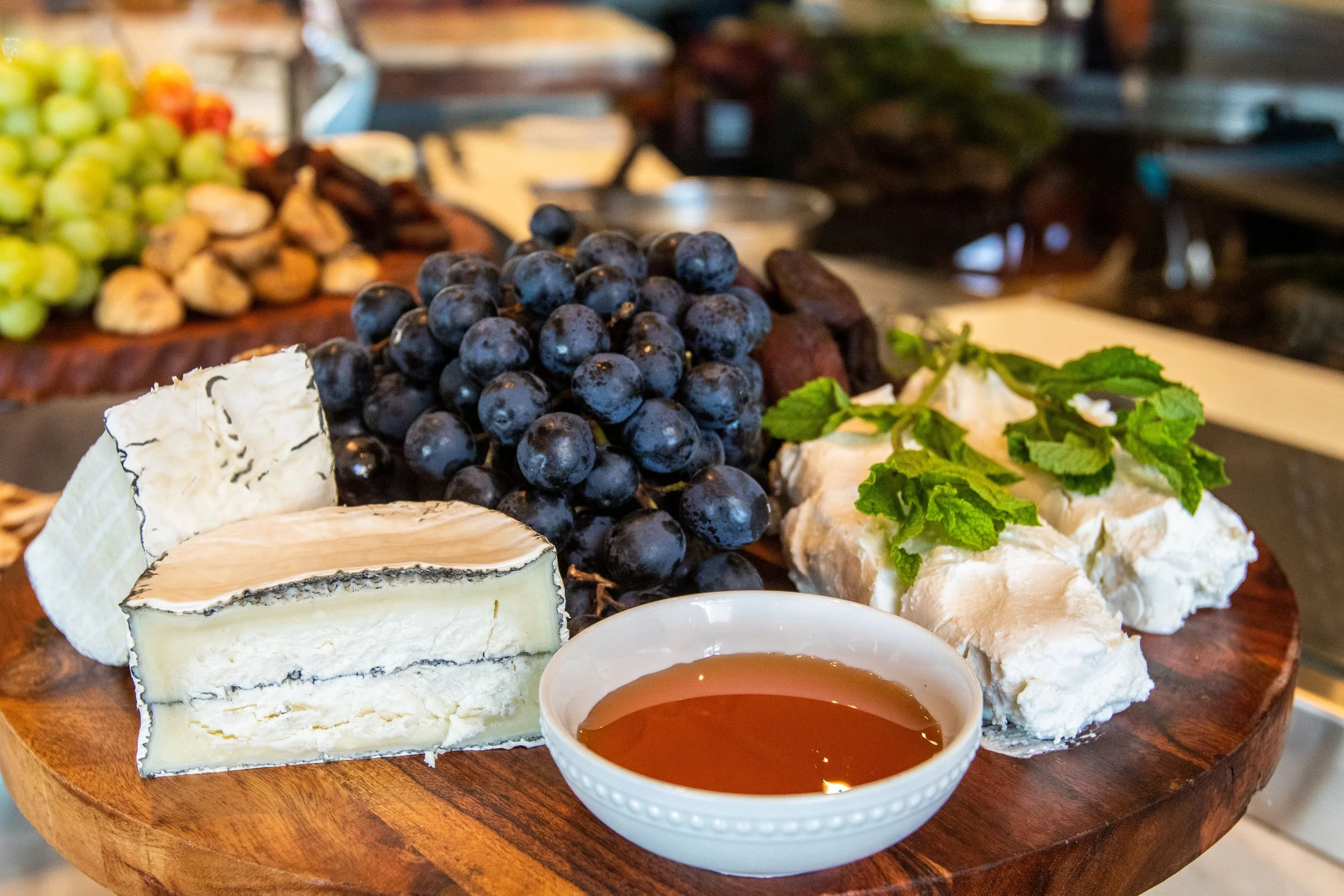 A cheese platter featuring various cheeses, purple grapes, and a bowl of honey, garnished with mint leaves on a wooden serving board.