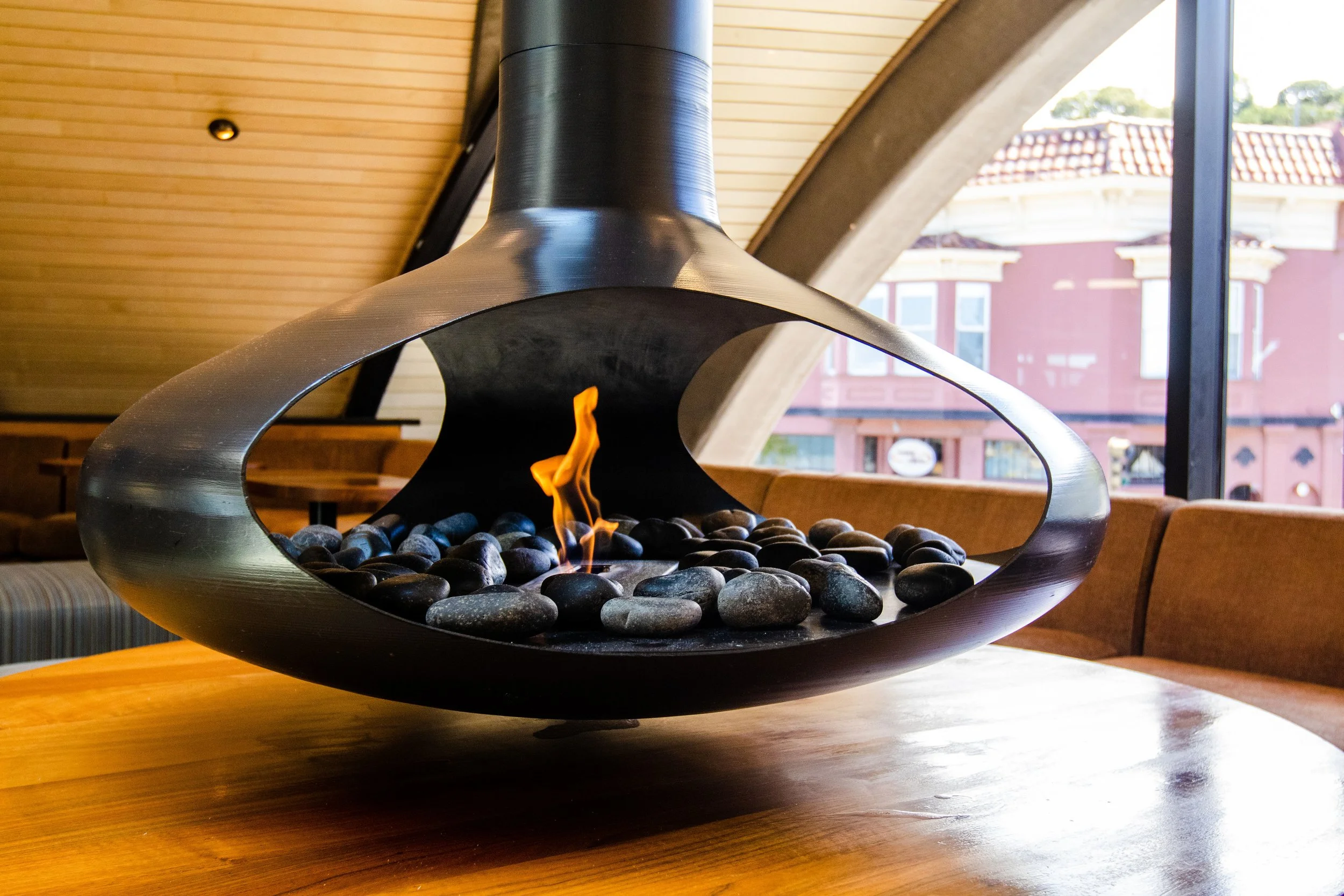 Modern tabletop fireplace with black stones and small flame in a restaurant or lounge setting with large windows and wooden ceiling.