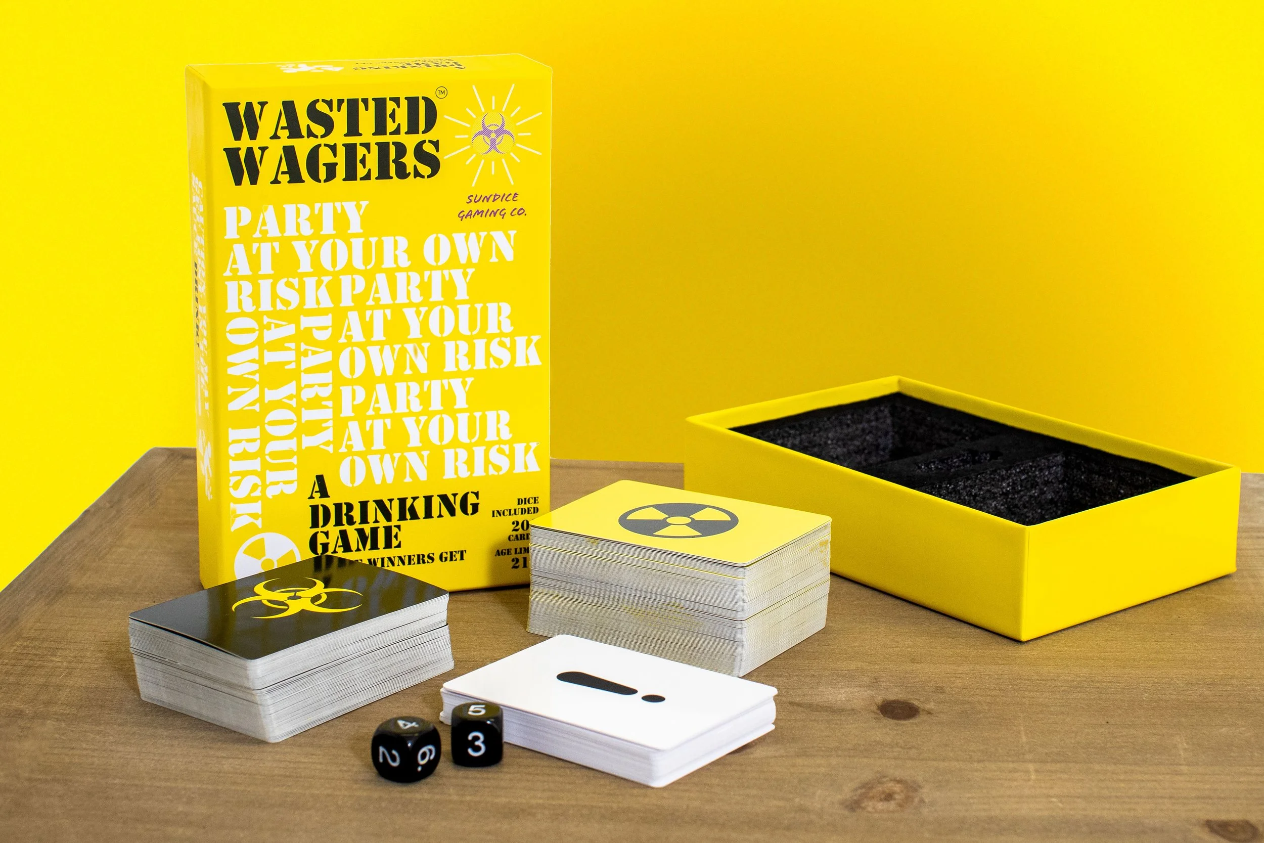 A card game called 'Wasted Wagers' with black and white cards, dice, and a yellow box on a wooden table against a yellow background.