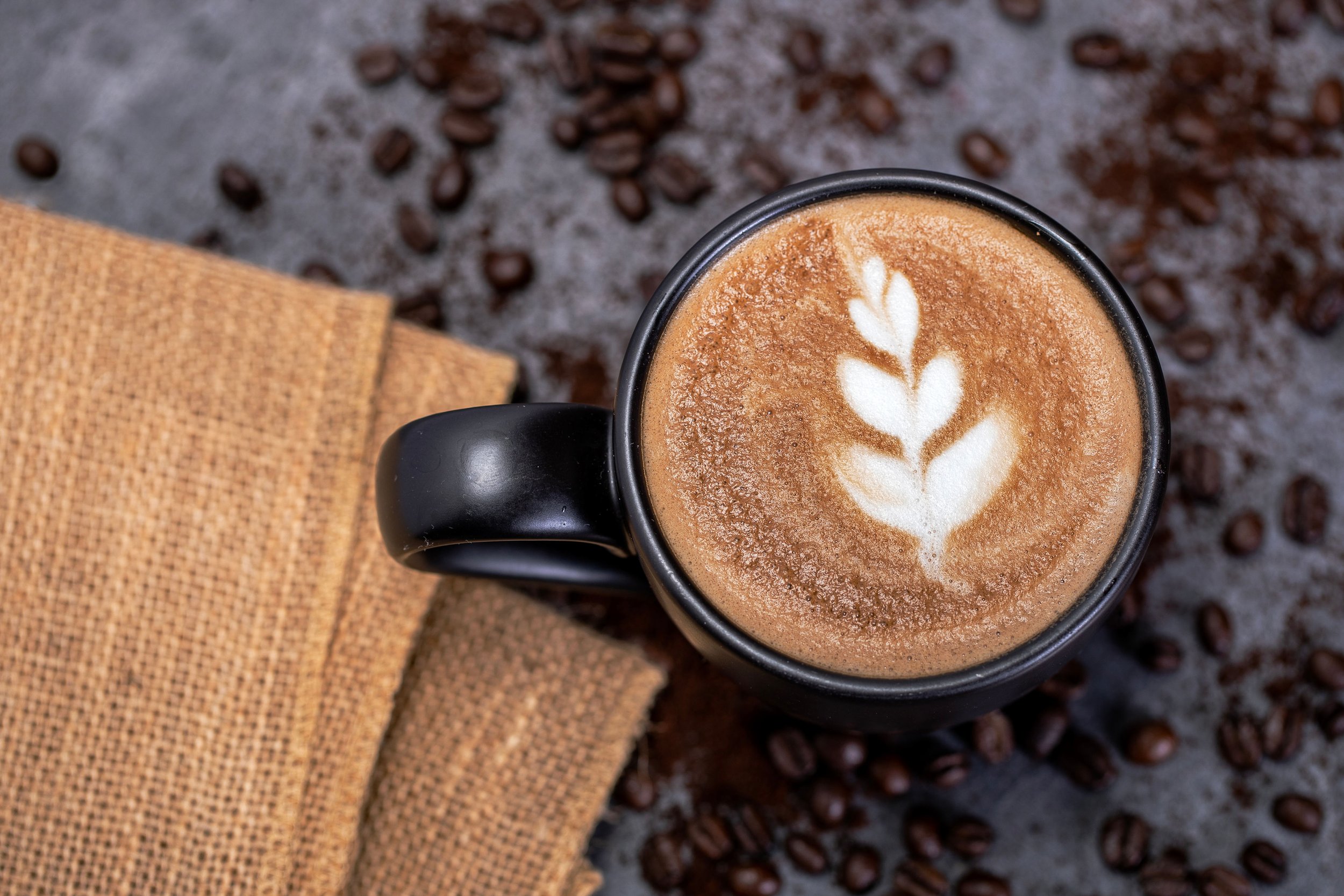 A black mug of latte with latte art on top, surrounded by coffee beans and a folded brown napkin.