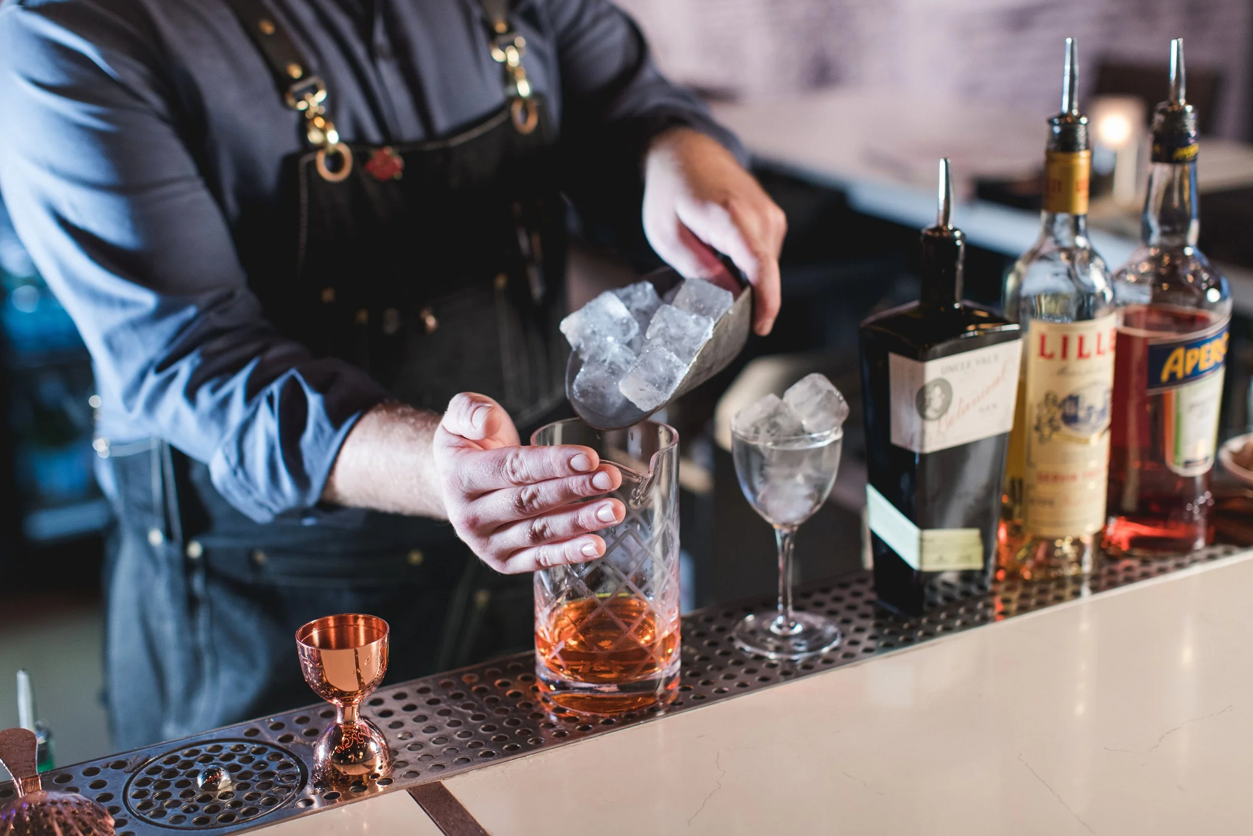 A bartender pouring a drink with ice into a glass at a bar, with bottles of alcohol and bar tools on the counter.