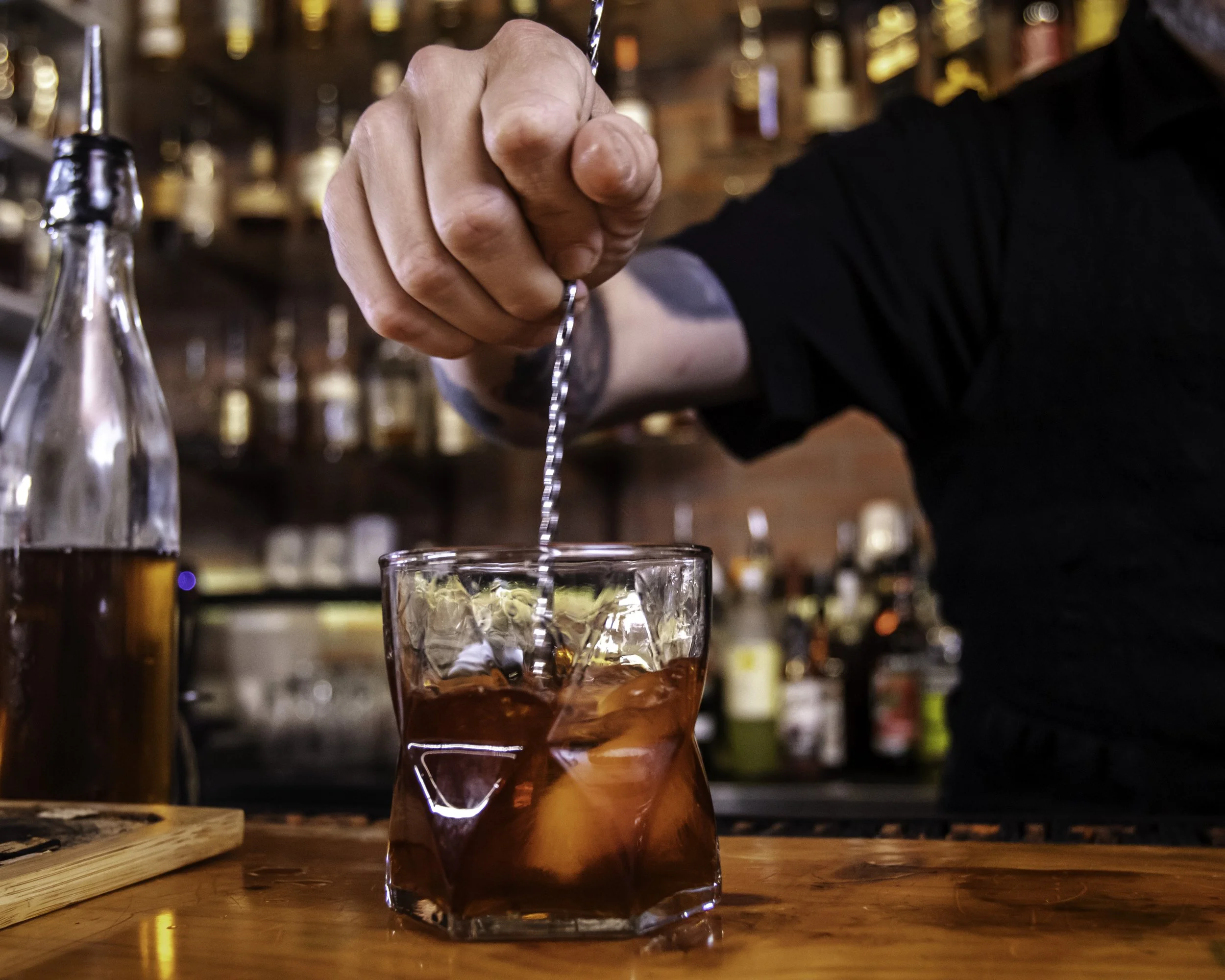 A bartender stirring a cocktail with a metal swizzle stick, with a glass of dark liquid and ice on a wooden bar counter, bottles of alcohol blurred in the background.