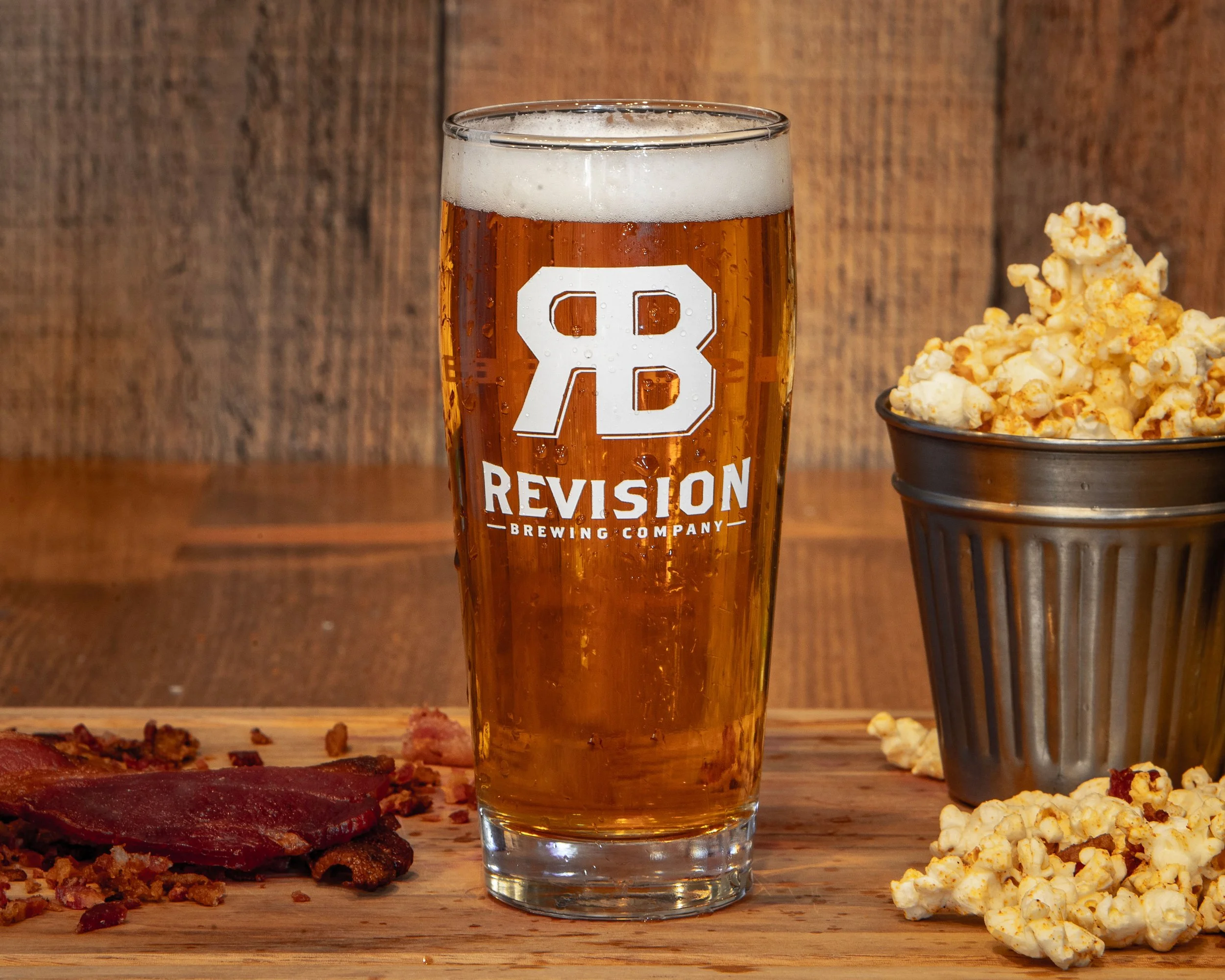 A glass of beer with the Revision Brewing Company logo, a bowl of popcorn, and slices of bacon on a wooden surface with a wooden wall background.