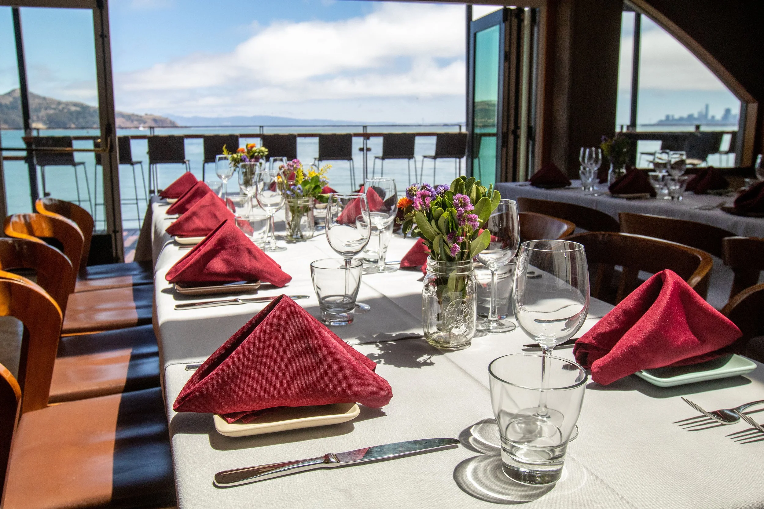 A restaurant table set for a meal with red napkins, wine glasses, water glasses, and a small floral centerpiece, overlooking a body of water and city skyline through open doors.