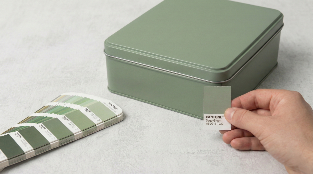Close-up of a rectangular sage green tin box with a matte finish, shown with a Pantone fan deck to demonstrate professional color matching capabilities by JB Packing Industrial.