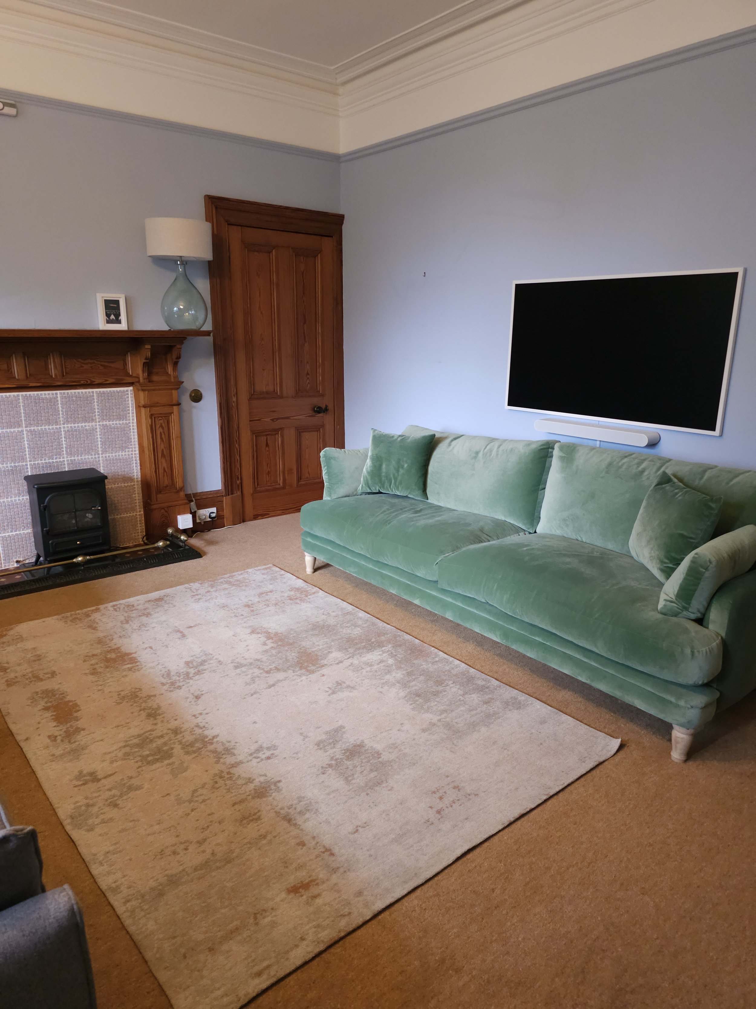 Living room with a green sofa, a rug, a wooden fireplace mantel, a mounted black television, a door, and a small lamp on the mantel.