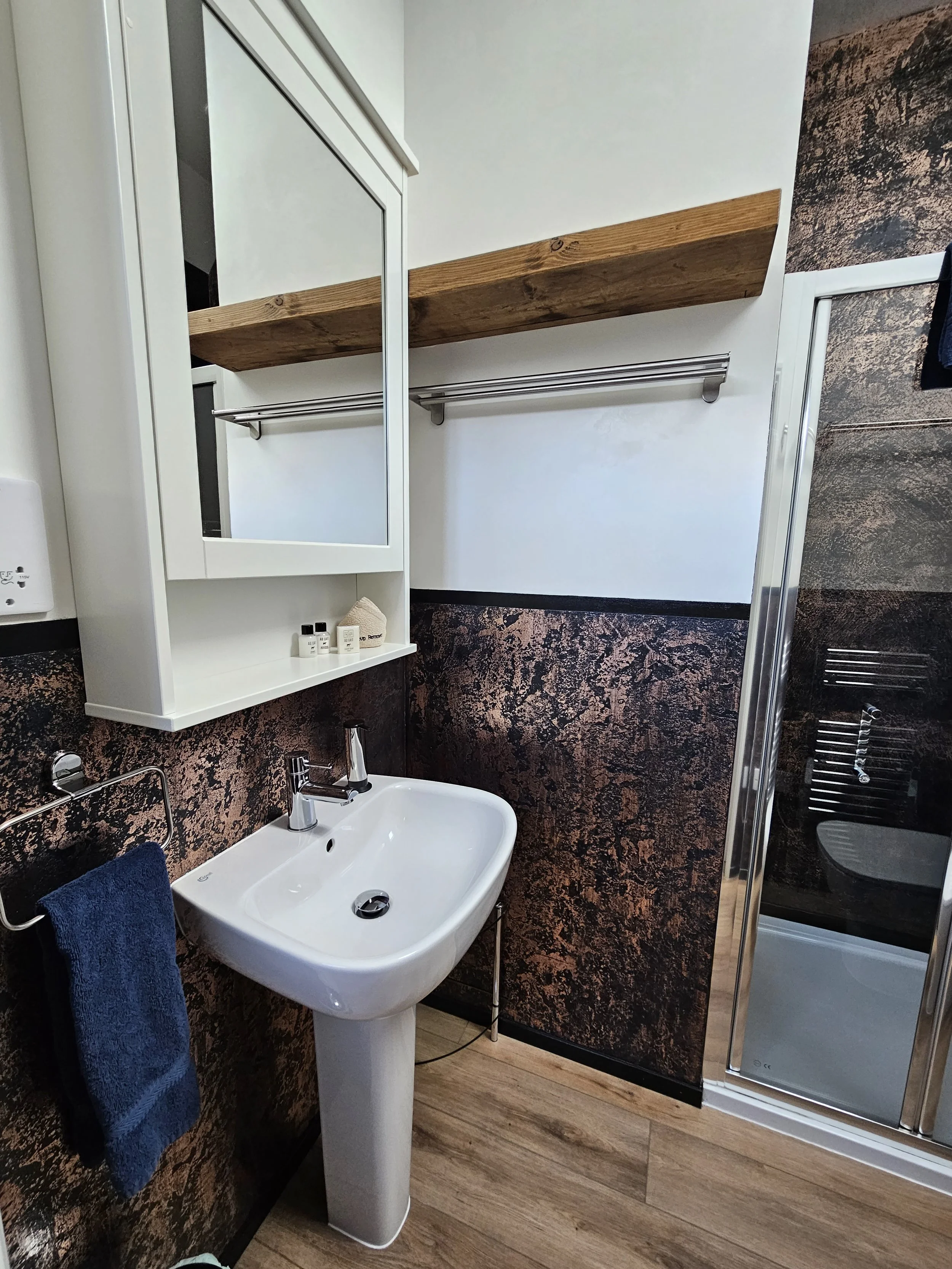 Small bathroom with a white sink and mirror, dark textured walls, wooden shelf, towel rack with a blue towel, and a glass shower door.