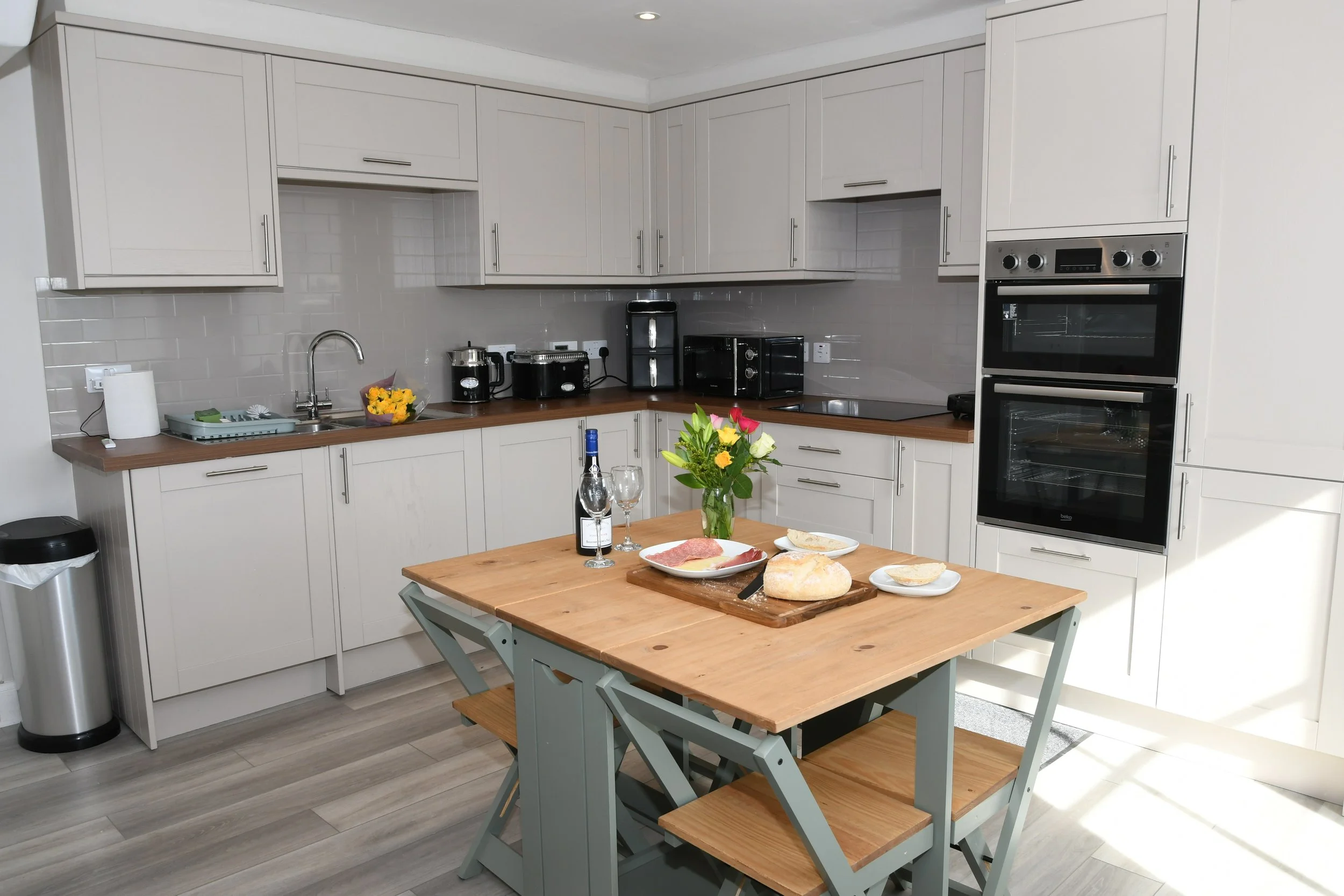 Kitchen with white cabinets, gray subway tile backsplash, and a wooden dining table set with bread, wine, and flowers. Holiday cottage in Newtonmore. Escape Cottages Scotland.