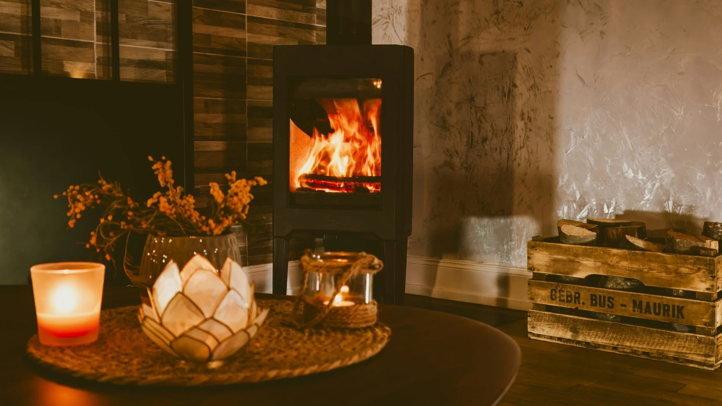 A cozy living room scene featuring a lit fireplace, a candle, a decorative flower container, and a glass jar with a candle on a round table. To the right, there is a wooden crate filled with cut logs. The room has warm lighting and textured walls.