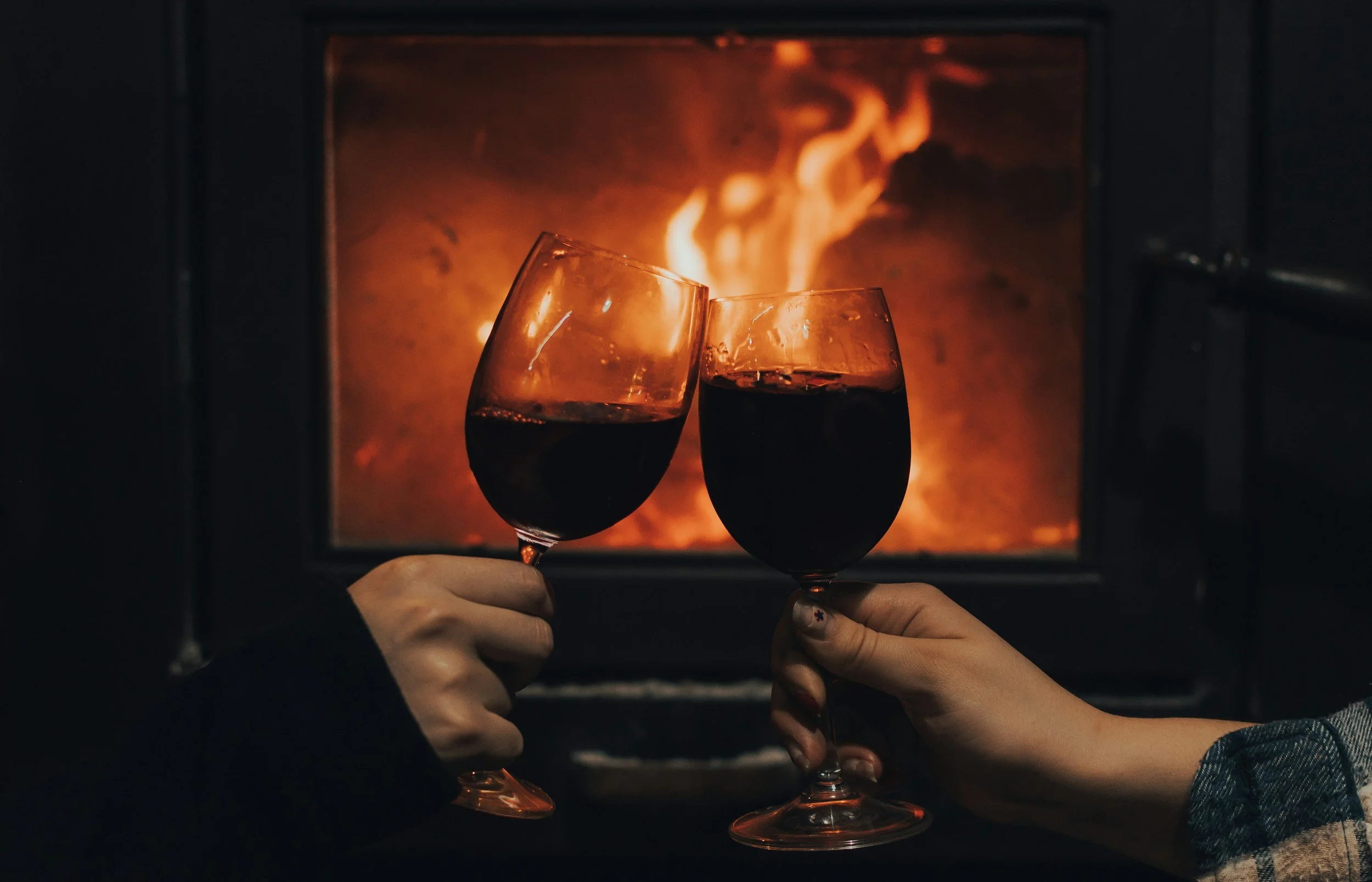 Two hands holding glasses of red wine in front of a lit fire in a fireplace.