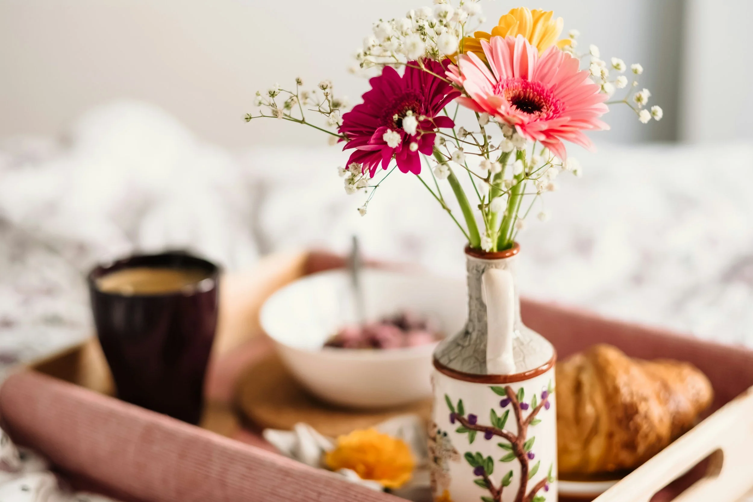 Close-up of a breakfast tray with a cup of coffee, a bowl of food, a piece of bread, and a ceramic vase with pink, yellow, and white flowers.