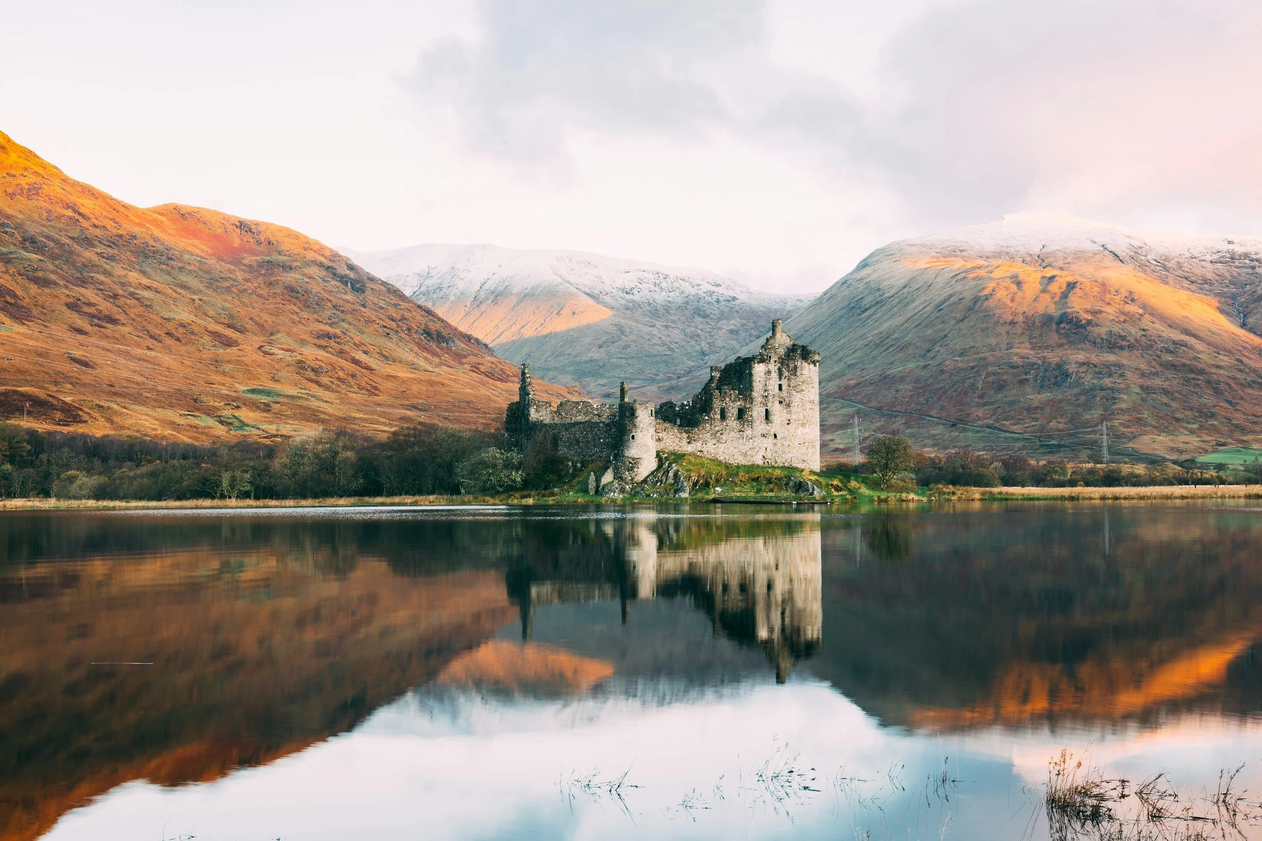 Ruined medieval castle on the edge of a calm lake with reflecting water, surrounded by hills with autumn foliage, under a cloudy sky.