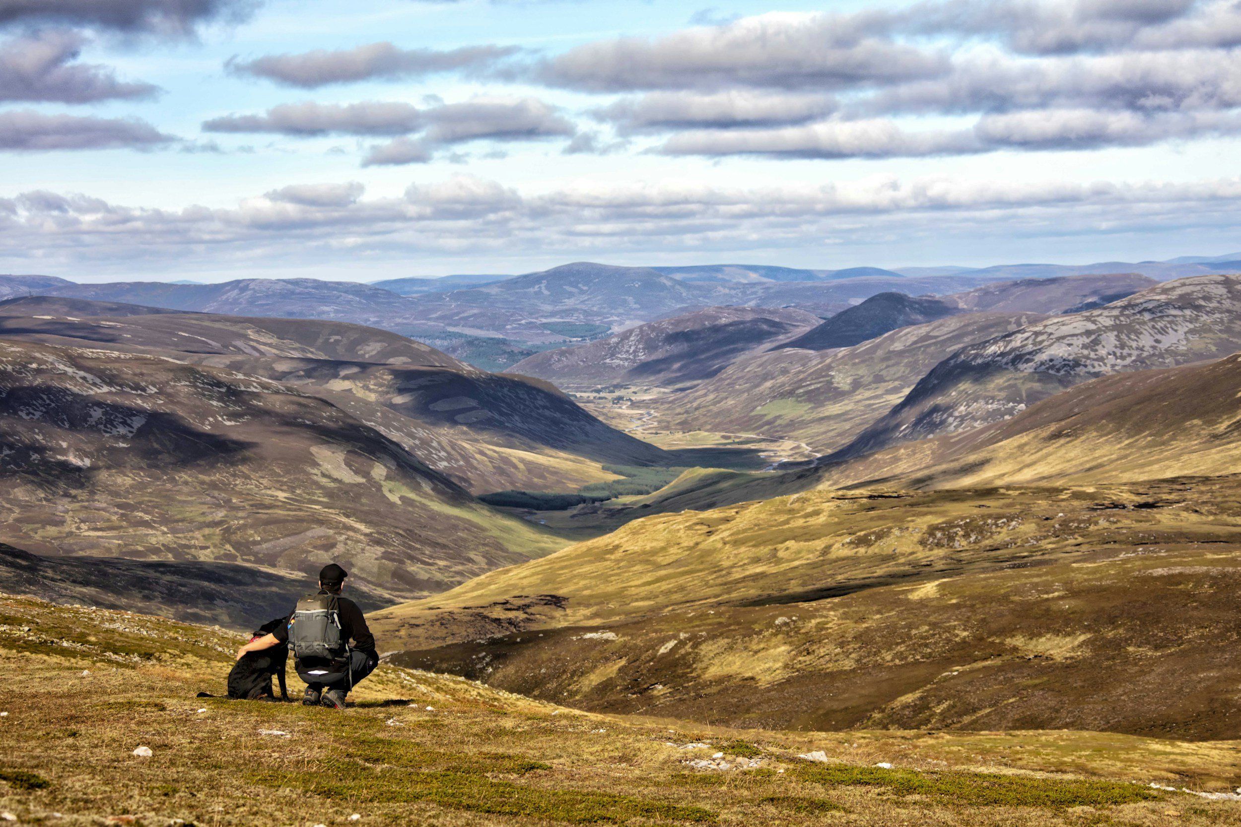 A person crouching on a grassy hilltop overlooking a vast valley surrounded by rolling hills and mountains, under a partly cloudy sky.