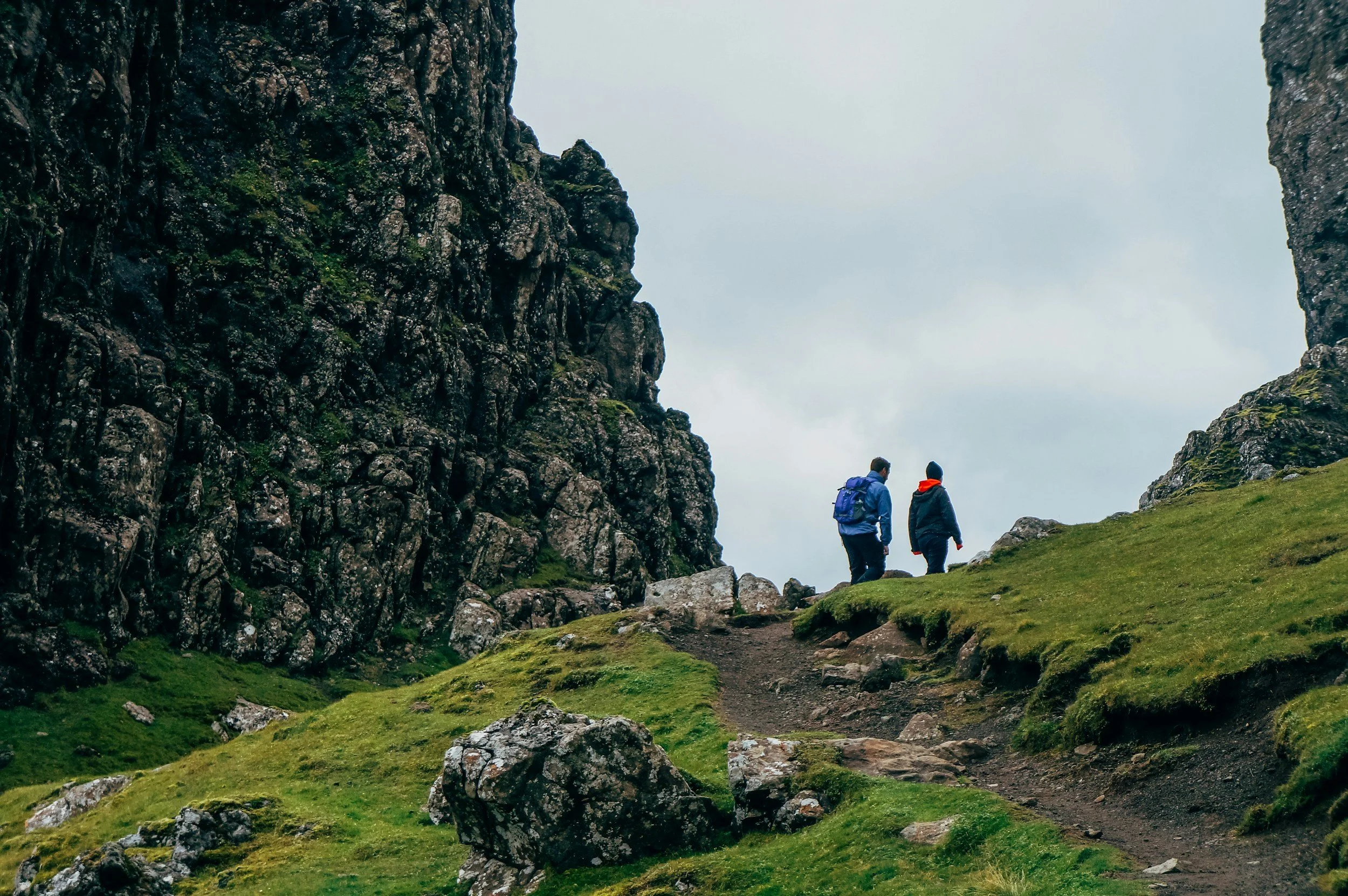 Two hikers with backpacks walking on a narrow trail between tall rocky cliffs and lush green grass under a cloudy sky.