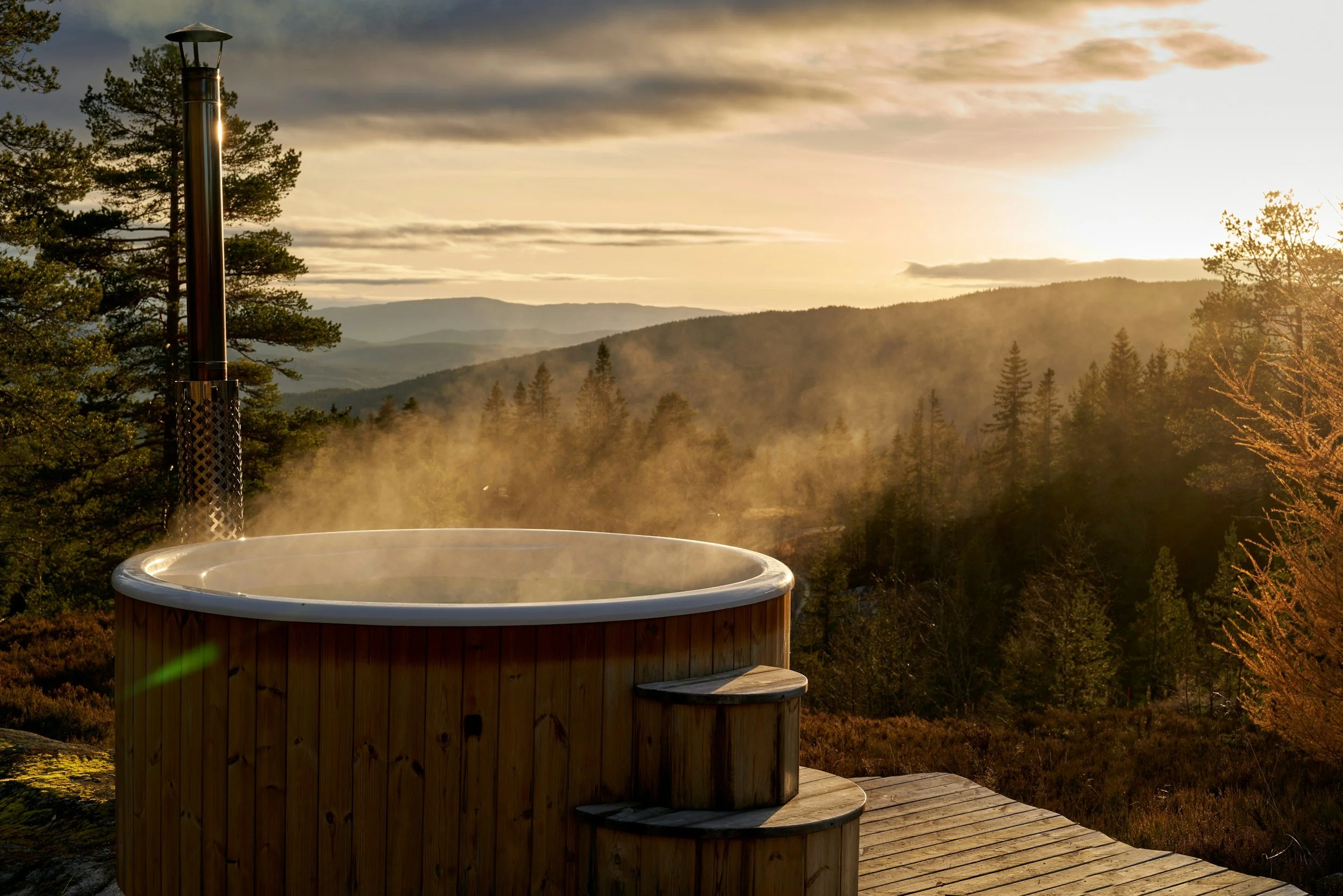 A wooden hot tub with steps in an outdoor setting, steam rising from the water, surrounded by trees and a forested mountain landscape at sunrise or sunset. - Escape Cottages Scotland