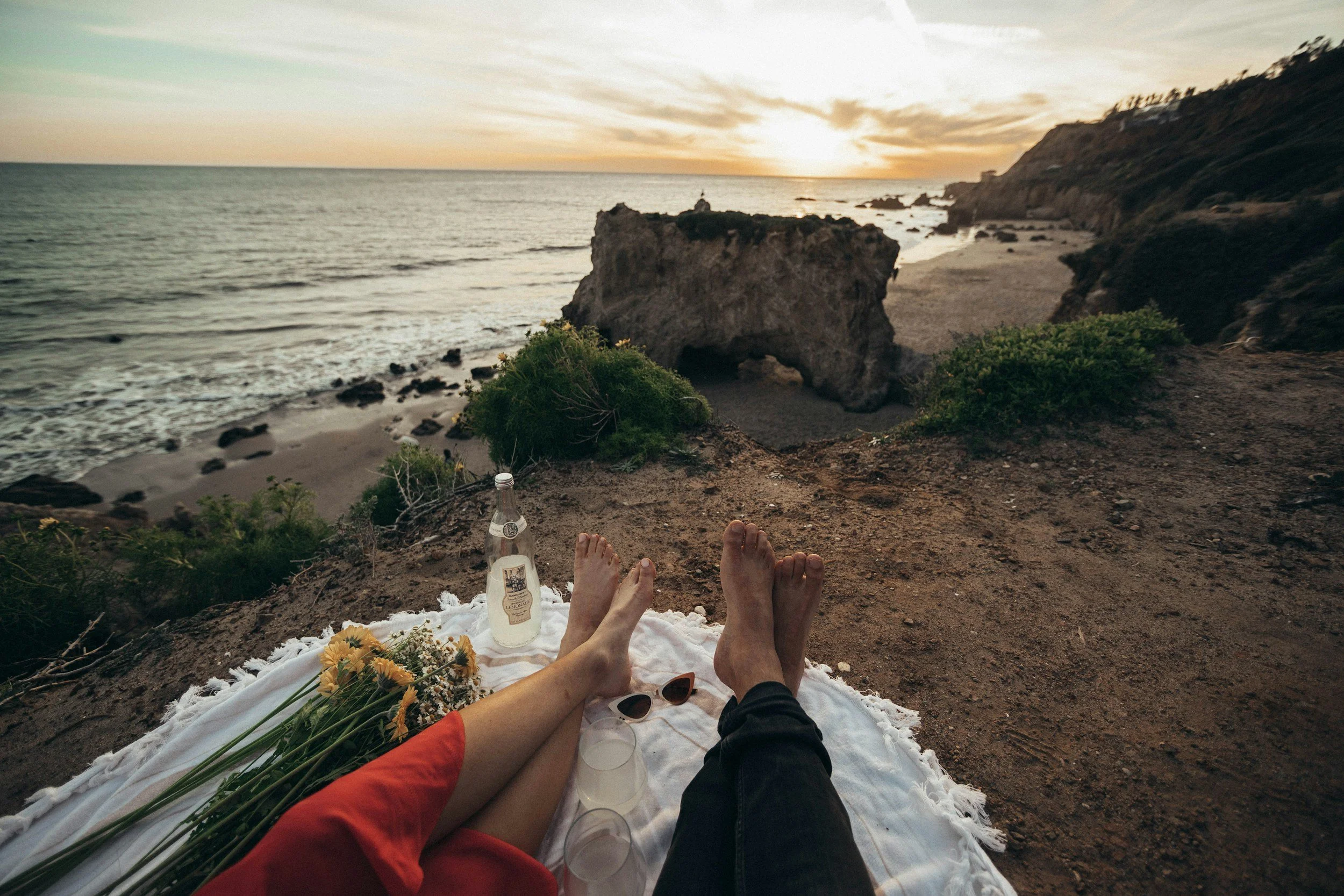 A person relaxing on a white blanket at the beach during sunset, with their feet visible, next to flowers, sunglasses, a bottle, and a paper cup; the background shows the ocean with waves and rocky cliffs.
