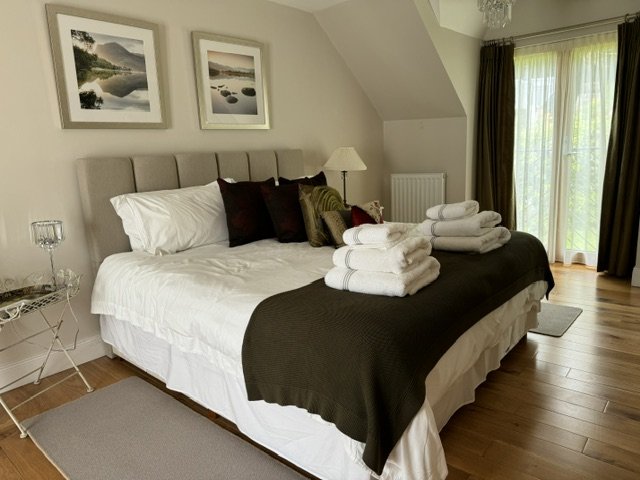 A neatly made bed with white sheets, dark brown throw blanket, and multiple pillows, topped with folded white towels, in a bright bedroom with hardwood floors, framed landscape art on the wall, and a sliding glass door with curtains leading outside.