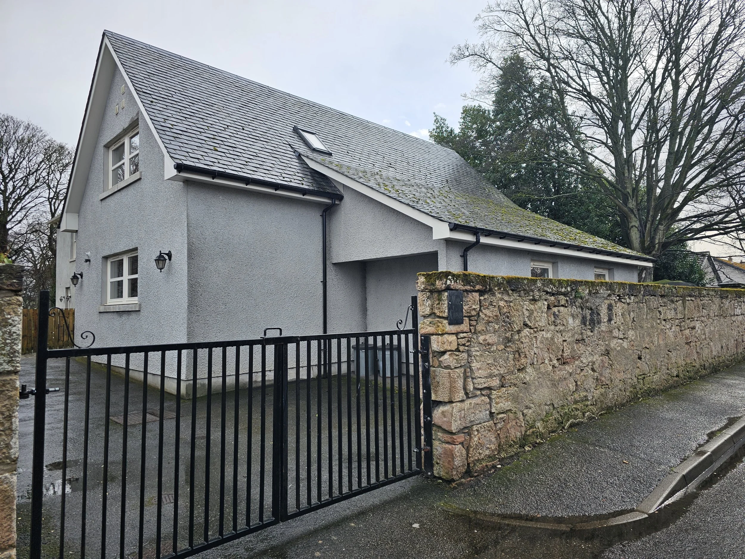 Wee Corsee, Nairn, Scotland with a gray exterior and black roof, surrounded by a stone wall and black metal gate, with a large tree in the background on a rainy day.