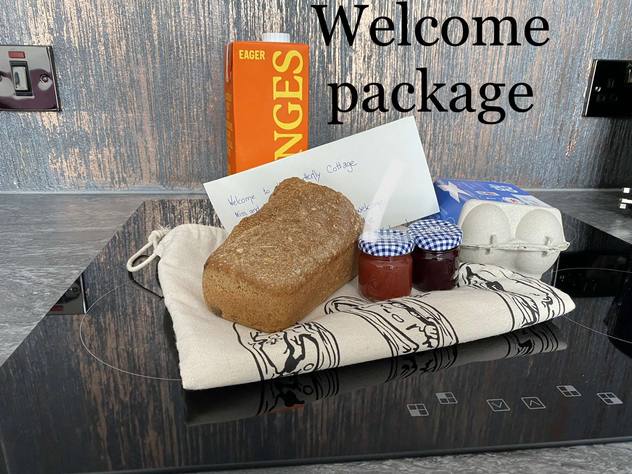 Welcome package on a black induction stove, includes a loaf of bread, two jars of jam with checkered lids, a carton of eggs, a note with a handwritten message, and a canvas bag, with a sign in the background that says 'Welcome package' and a box of Eager Snanges on the wall.