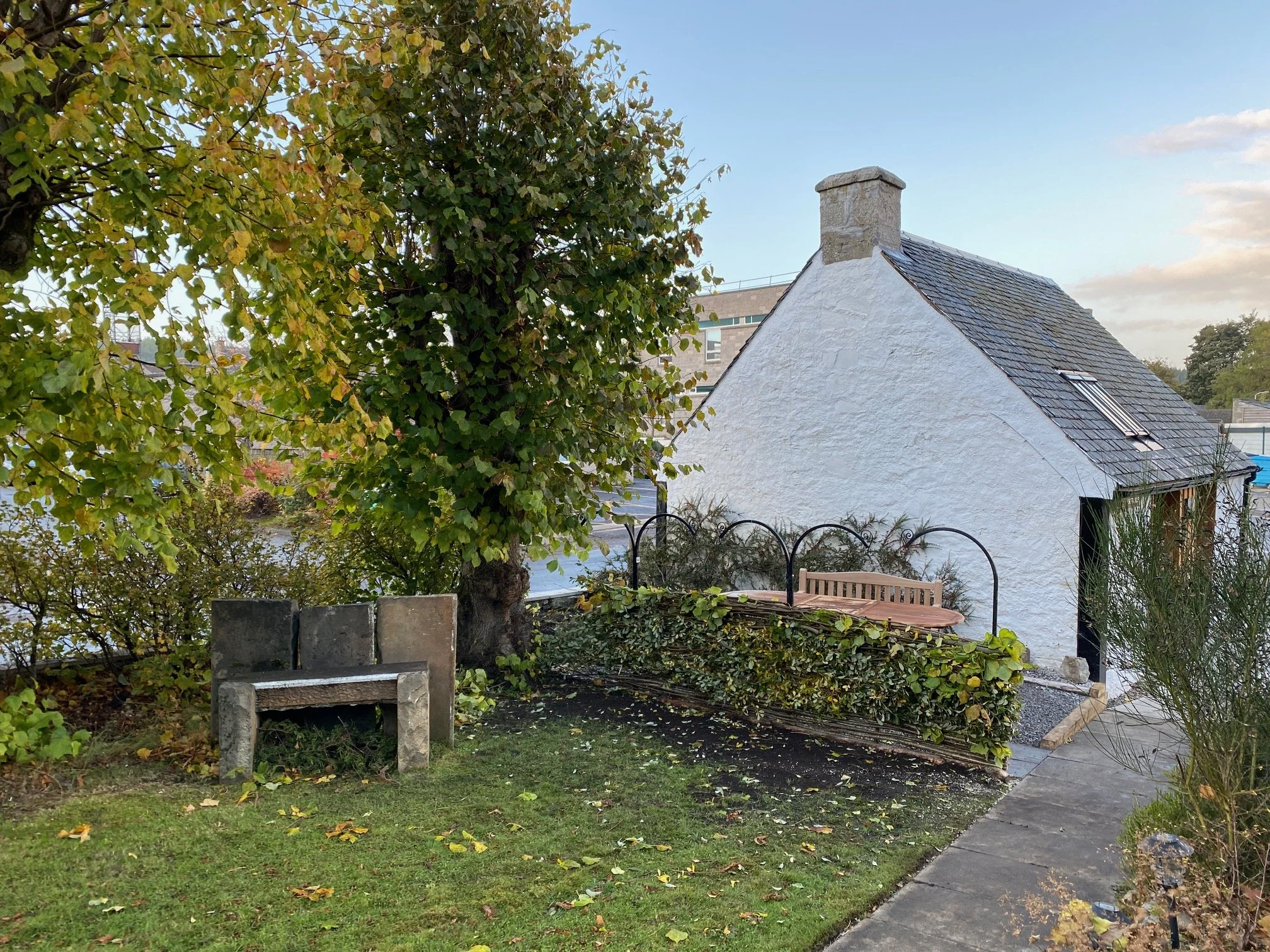 Small white house with a sloped roof, chimney, and a skylight, surrounded by trees and a garden with a stone path, showing a wooden bench and trellises, during daytime.