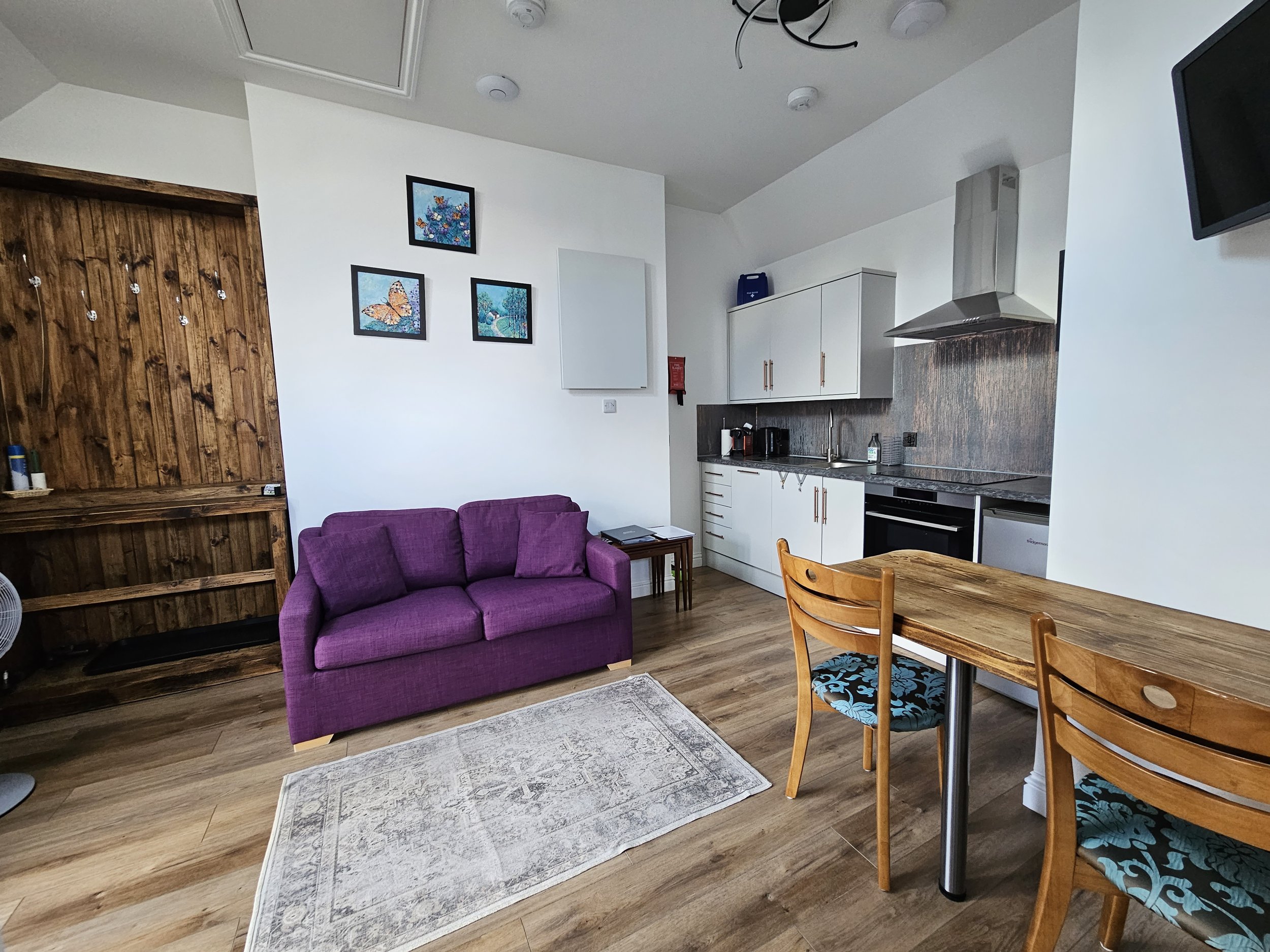 Interior view of a living area with a purple sofa, a small wooden dining table with two chairs, a kitchen with white cabinets and stainless steel appliances, and a wall decorated with three framed pictures of butterflies and nature scenes.