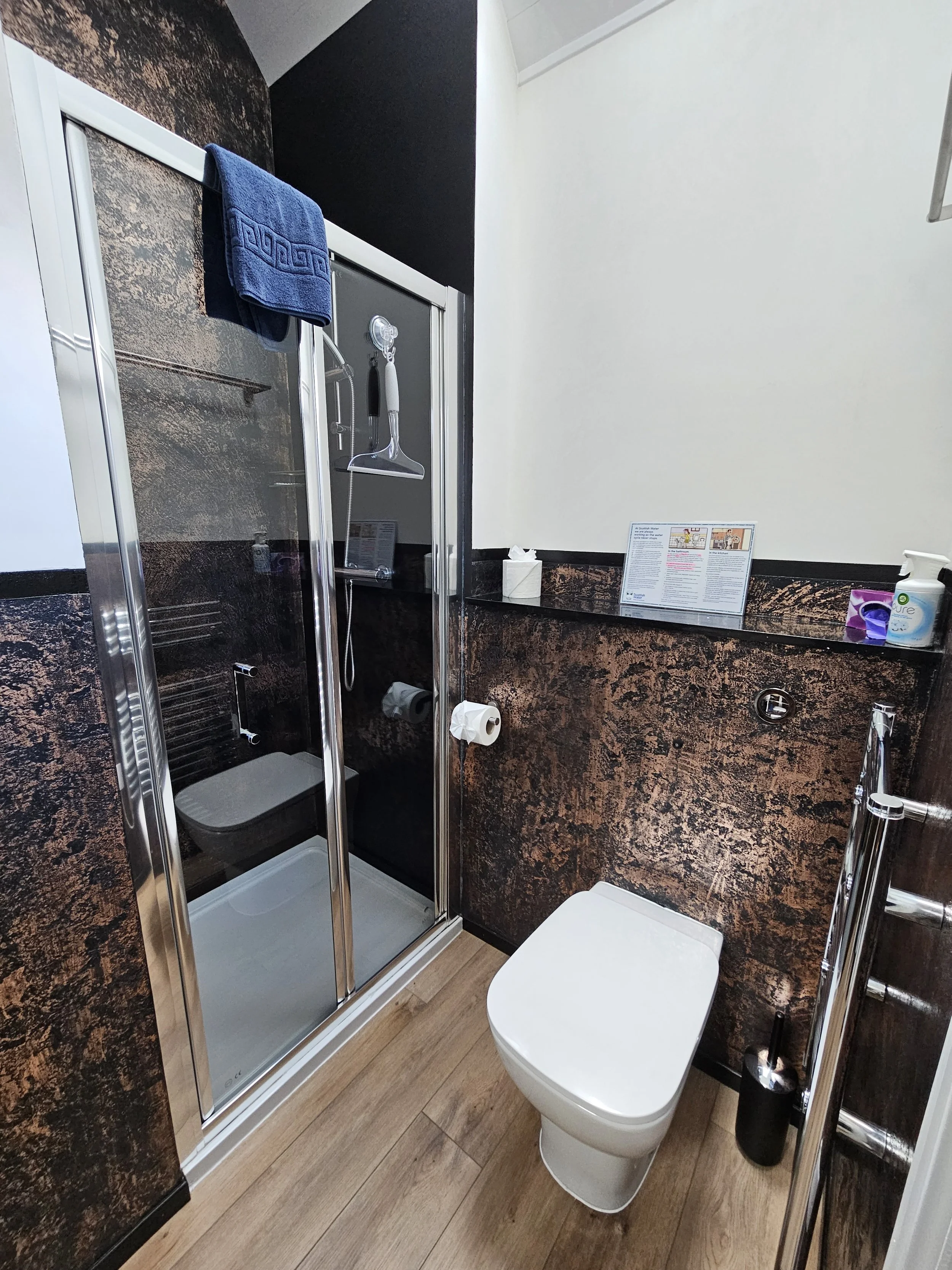 A small bathroom with a glass-enclosed shower stall, a white toilet, a wood-patterned vinyl floor, and dark brown and white walls. A blue towel hangs on a towel rod above the shower, and there are toiletries and a paper towel roll on a ledge above the toilet.