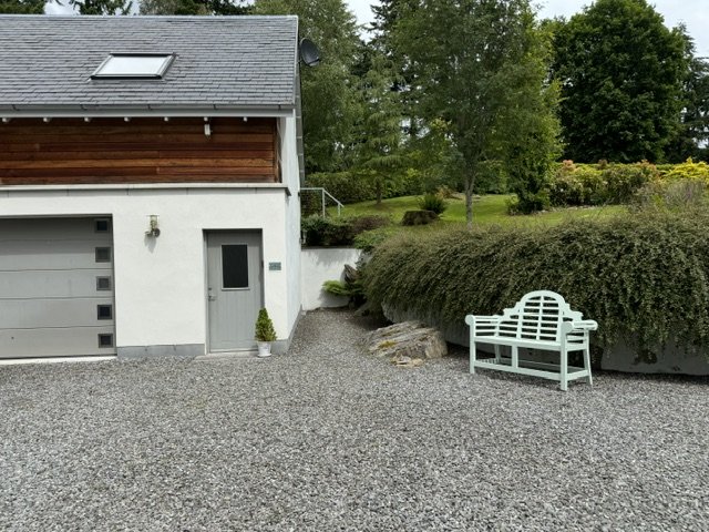 Residential house with garage, side door, gravel driveway, white bench, and landscaped yard with bushes and trees.