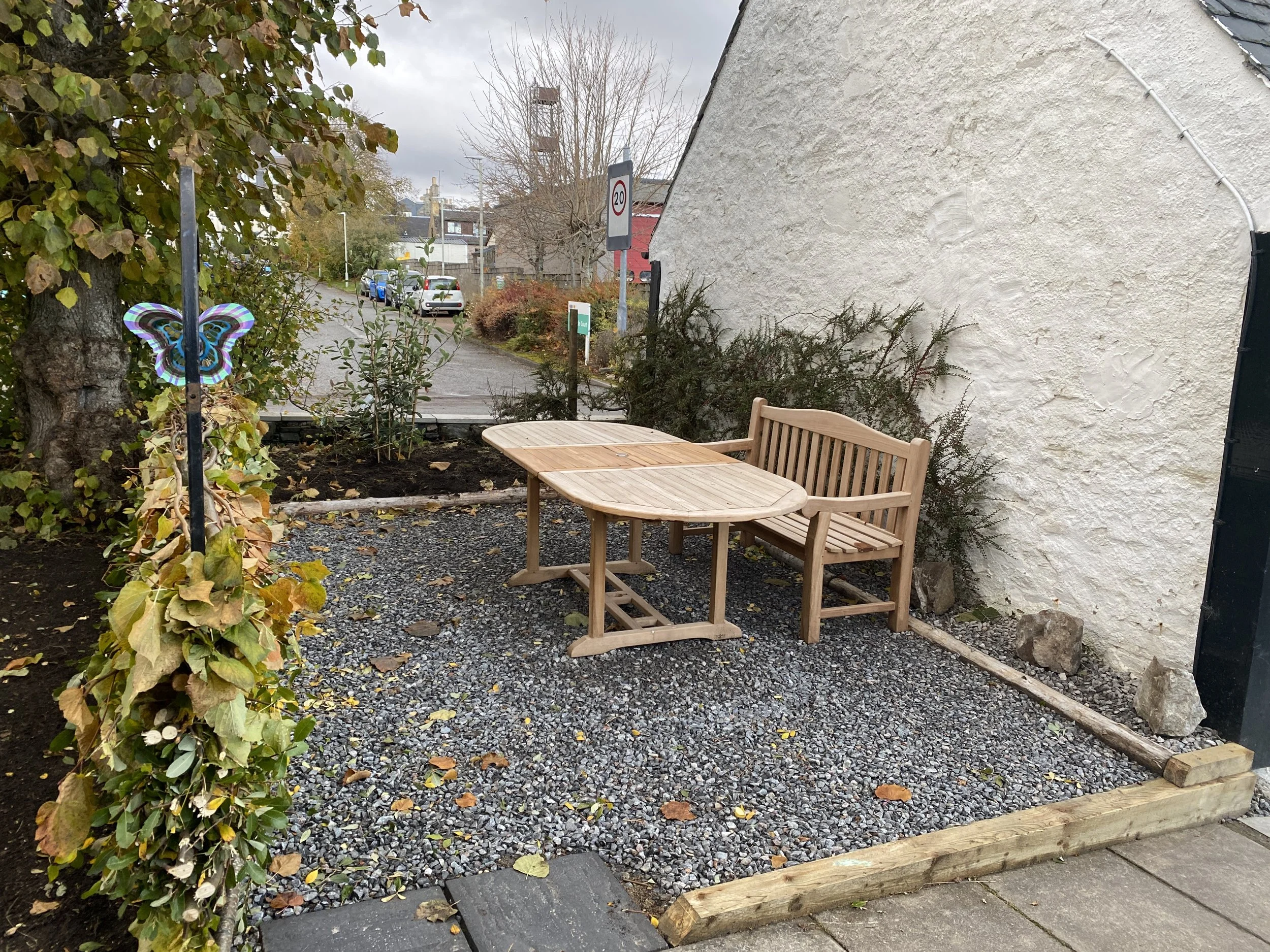 A small outdoor seating area with a wooden bench and a matching table on a gravel surface, surrounded by plants and trees, next to a white stucco wall with an electric conduit, on a cloudy day.