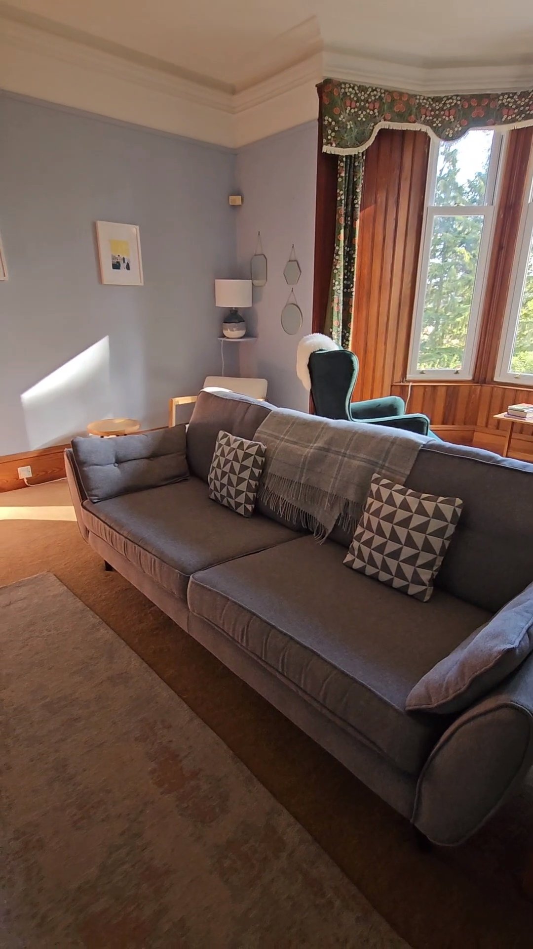 A cozy living room with a gray sofa, patterned throw pillows, and a blanket on top. Behind the sofa is a window with floral curtains and wooden paneling, letting in natural light. There is a green armchair near the window, and a small side table with books. The wall has framed artwork, a wall lamp, and decorative mirrors.