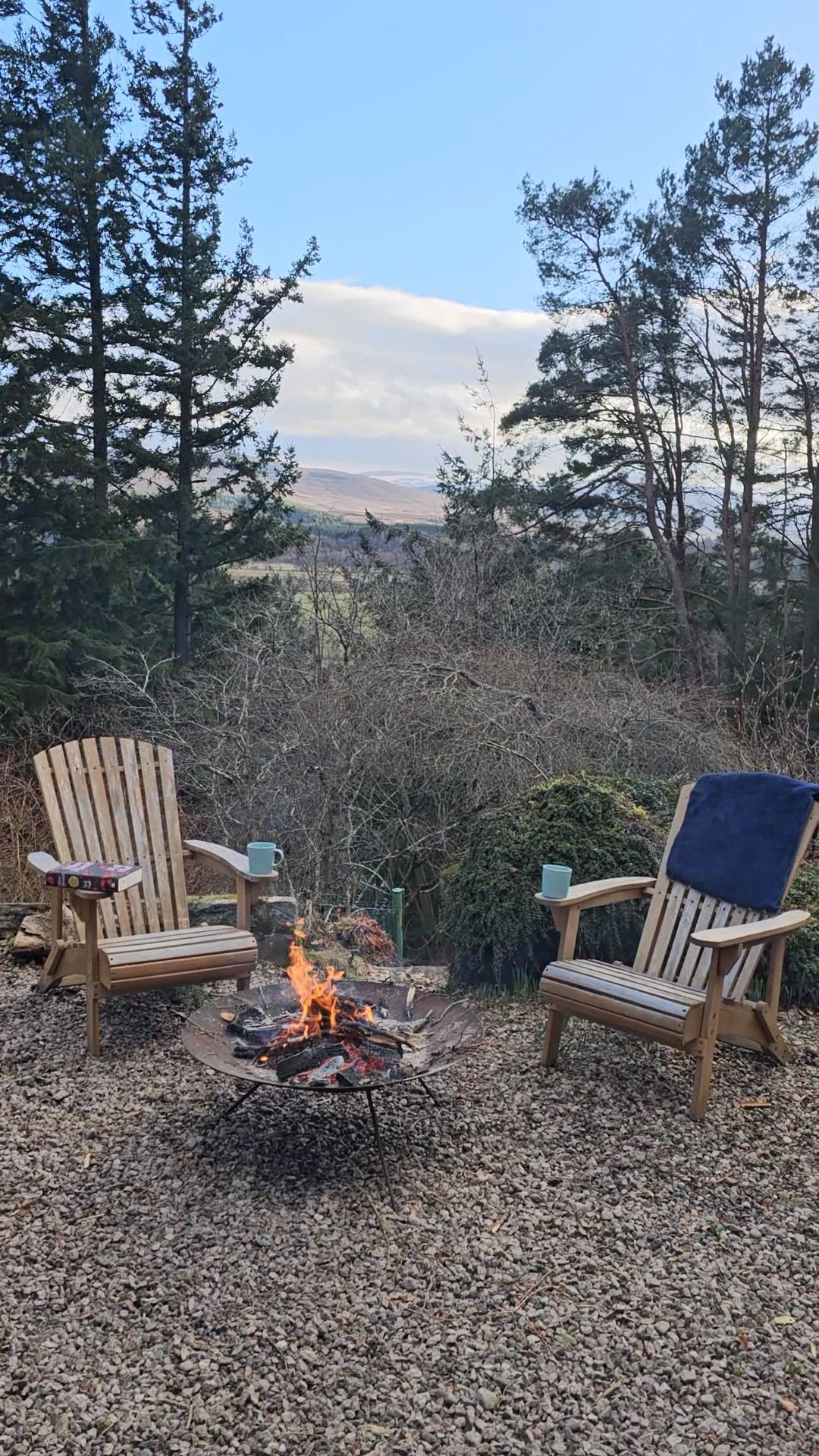 Two wooden chairs around a small fire pit on a gravel surface, overlooking a wooded landscape with trees, bushes, and distant hills.